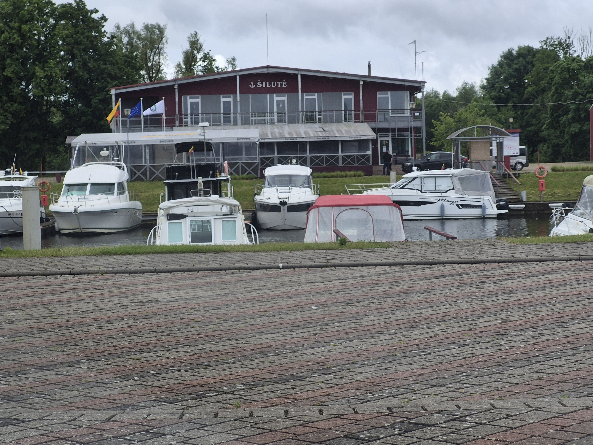 The image depicts a waterfront scene in Šilutė, Lithuania, likely at a marina or harbor. In the foreground, a paved area with diamond-patterned concrete blocks forms the ground. Beyond this, a body of water, likely a river or bay, is visible, lined with green grass. Moored boats of various sizes are present on the water.
Prominently featured is a two-story building with a red facade and gray trim, labeled "ŠILUTĖ" in white lettering. This building appears to be a restaurant, club, or public facility, with large windows and balconies. Flags, including the Lithuanian flag and the European Union flag, are displayed on poles in front of the building.
Several boats are docked. On the left, a white motor yacht is moored. Next to it, another white boat with a covered cabin is visible. A white boat with a red canopy is in the center, and to its right, a larger white motor yacht with the name "Maya" on its side is docked. Further to the right, another white boat with a canopy is partially visible. A dark-colored car is parked near the building on the right side of the image.
The environment is overcast, suggesting a cloudy day. Trees and vegetation are visible behind the building and on the surrounding land. The scene conveys a sense of a recreational area by the water.