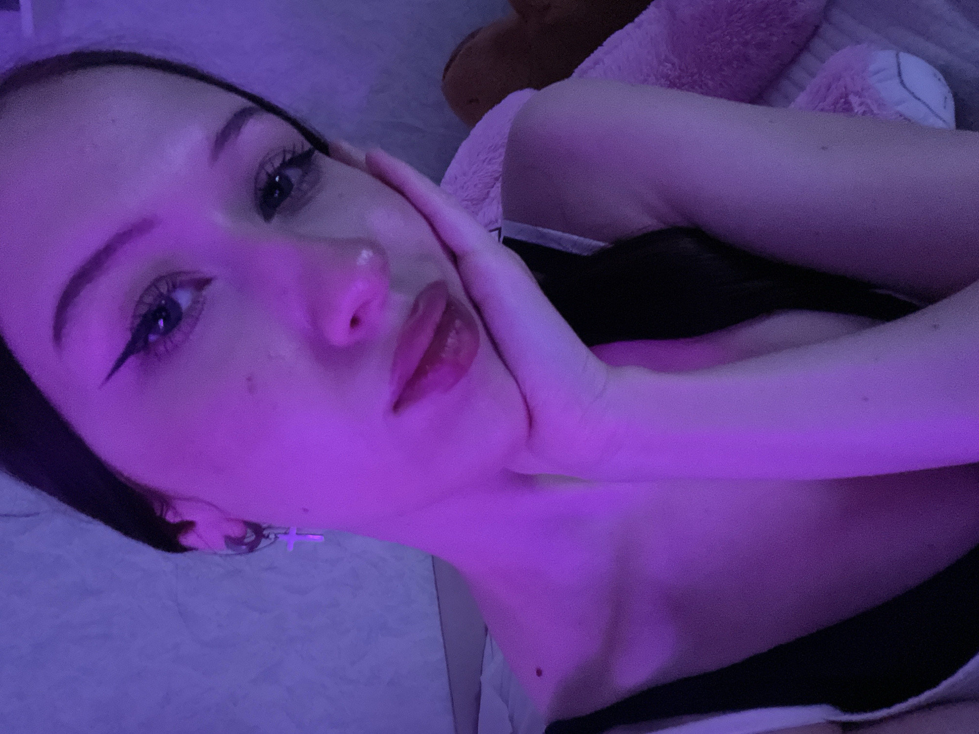 This close-up portrait, set in the city of Votkinsk, Russia, features a young woman bathed in pervasive purple/magenta ambient light. She has long, dark hair, partially visible, and is wearing a dark top with white straps. Her make-up includes prominent black winged eyeliner and a shiny lip product. A silver-toned hoop earring with a dangling cross charm is visible in her left ear. She is posed with her right hand supporting her chin and lower jaw, fingers lightly curled. Her gaze is directed towards the viewer with a neutral to slight smile. A dark mole is present on her left collarbone area. The background consists of a light-colored, textured wall and what appears to be a light pink, fluffy pillow or stuffed animal on the right, suggesting an indoor setting, likely a bedroom.