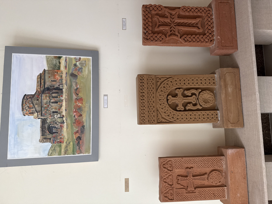 An indoor scene displays cultural artifacts against a plain light-colored wall, likely within a museum or exhibition space in Talin, Armenia. Centrally positioned on the wall is a framed painting, depicting the ancient, partially ruined Talin Cathedral, an edifice of dark stone with a central dome, set amidst a sparse landscape with scattered reddish and gray rocks under a light blue sky. Below this painting, three carved stone monuments, identified as khachkars (Armenian cross-stones), are exhibited on a light-colored plinth. The leftmost and rightmost khachkars are rectangular and reddish-brown, each featuring a prominent ornate cross with a circular element at its base, encased within decorative borders. The central khachkar is taller and lighter in tone, displaying an elaborate cross with swirling motifs and a circular base, enclosed within an intricately carved arched frame with braided patterns. Small white labels with black text are affixed to the wall below the painting, two of which partially read "TALIN...", confirming the geographical context. No individuals are present in the scene.