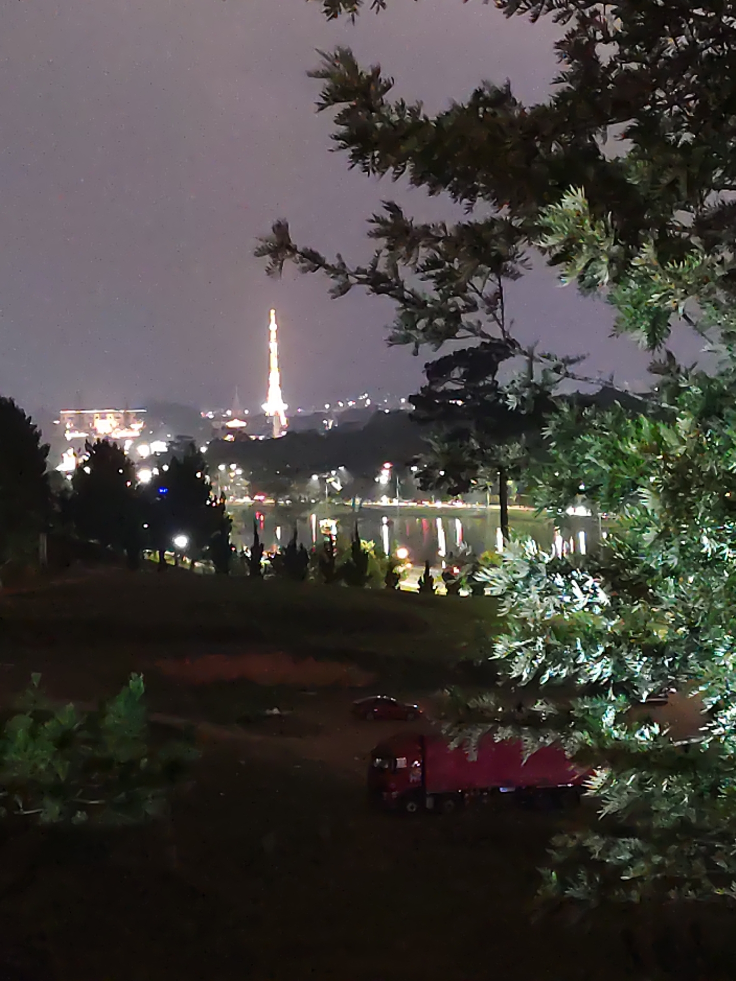 The image presents a night-time view of a cityscape from an elevated perspective, partially obscured by trees in the foreground. In the background, a illuminated tower stands prominently, its light accentuating its height against the dark sky. Adjacent to the tower, buildings with numerous lights contribute to the urban glow. A body of water reflects the city lights, creating a luminous streak across the center of the frame. In the lower part of the picture, a red truck is visible, parked on a dirt surface. The surrounding area includes dark, grassy terrain. The location is Lâm Viên Square in Đà Lạt, Vietnam.