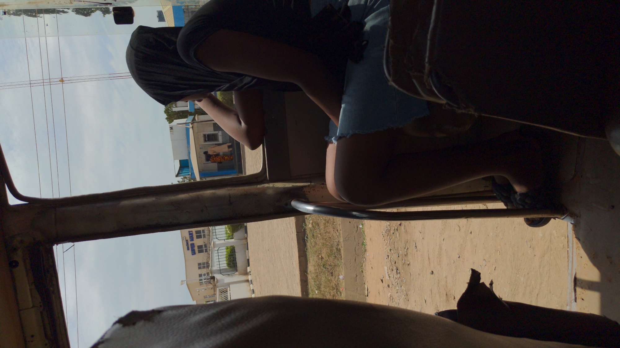 A view from inside a vehicle looking outwards, showing a person wearing a dark shirt and denim shorts. The person's face is covered. The vehicle's frame and parts of the interior are visible. Outside, there's a building labeled "SUPER MART" and another one with a partially visible sign, along with a street. The environment appears to be Ado, Nigeria.