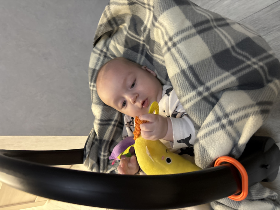 A baby, swaddled in a gray and white plaid blanket, sits in a black baby seat. The child is holding a yellow banana-shaped toy with purple and orange accents. The baby wears a white shirt with black whale silhouettes. The seat's frame, with an orange handle, is visible in the foreground. The background features a white surface, possibly a floor, and a gray wall.