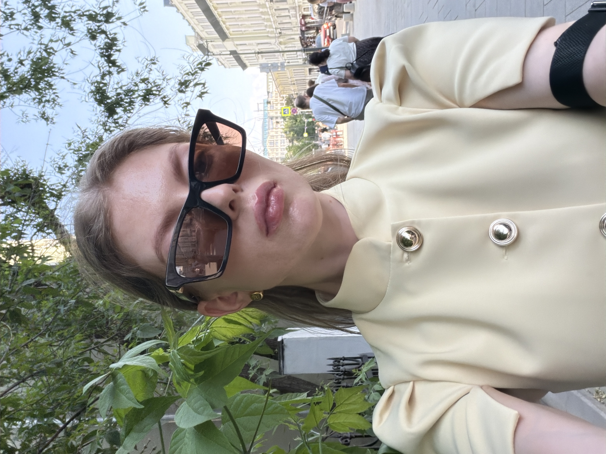 The image depicts a person wearing sunglasses and a light yellow shirt with gold buttons, with greenery in the foreground. In the background, a Moscow street scene is visible, featuring a building with white facade, people walking on the sidewalk, and street signs indicating it is likely a public area. A watch is present on the person's wrist.