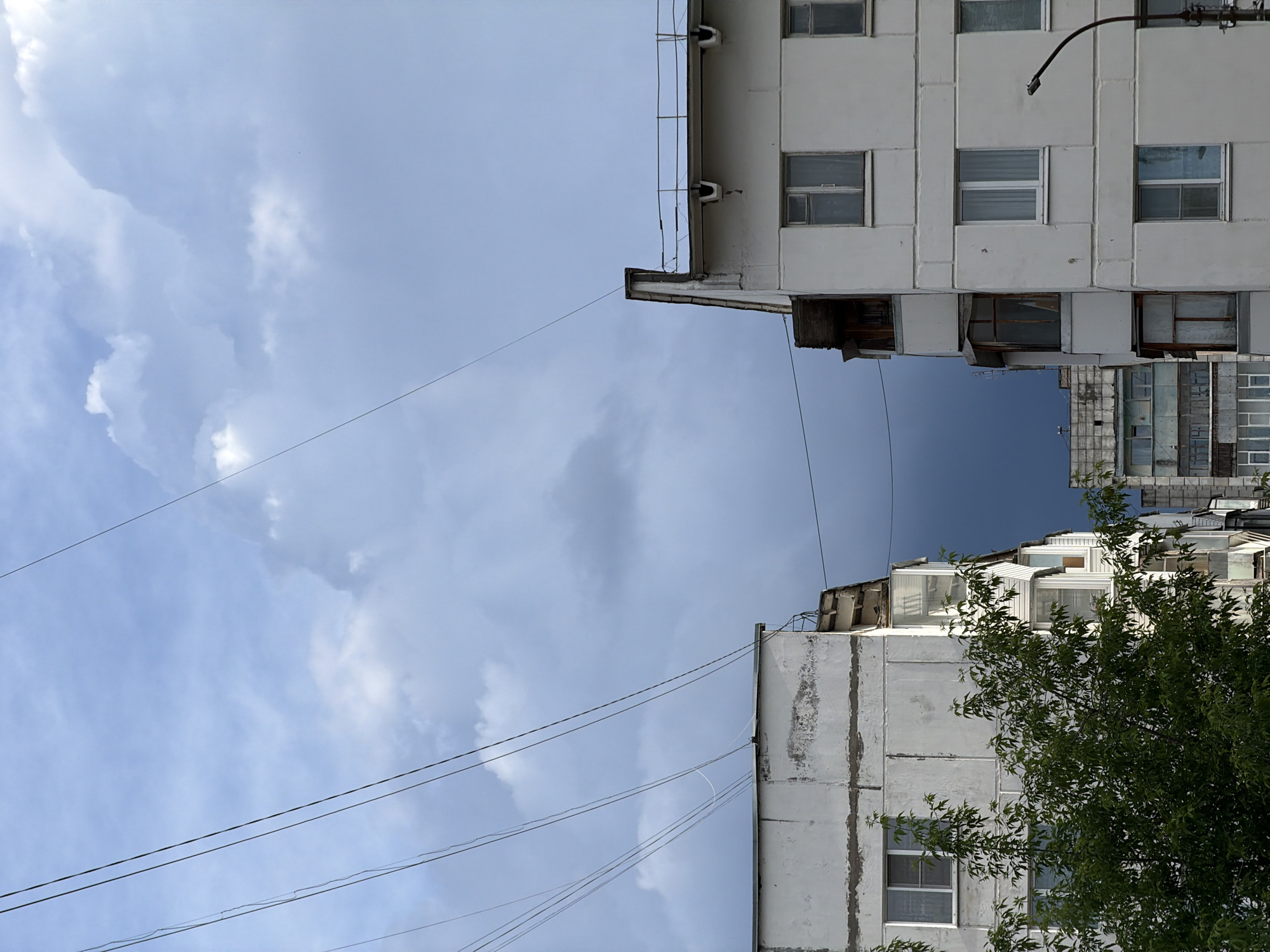 The image depicts an upward view of two buildings in what appears to be a Russian city, potentially Chaikovsky. The buildings are constructed of a light-colored material, possibly concrete or stucco, and exhibit a rectangular, block-like design with rows of windows. Some windows have visible curtains or blinds. There is evidence of aging and weathering on the building exteriors, including discoloration and patches. The buildings are connected by thin electrical wires that span across the open space between them. A portion of a tree with green foliage is visible on the right side of the frame, partially obscuring one of the buildings. The sky fills the majority of the image, exhibiting a mix of blue and white clouds, suggesting either partly cloudy or overcast conditions. The image is shot from a low angle, emphasizing the verticality of the buildings and the expanse of the sky. There is a dark blue section in the center above the buildings.