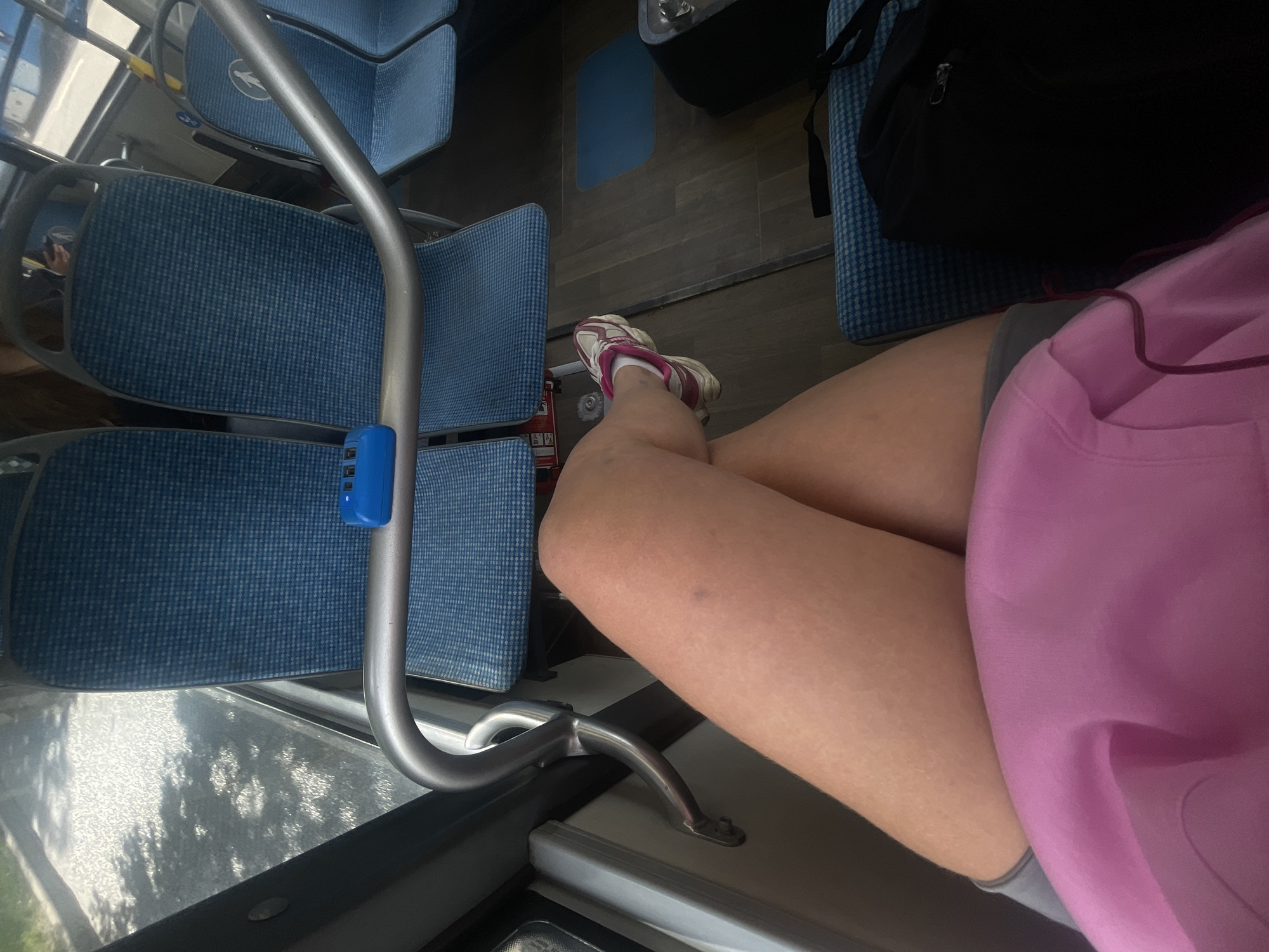 The image depicts the interior of a bus, possibly in Moscow, Russia, based on the Cyrillic characters on the blue box attached to the handrail. The focus is on the lower half of a person sitting on the bus. The person is wearing pink and grey athletic clothing. One foot, clad in a white and burgundy sneaker, is visible, resting towards the middle of the bus floor. A black backpack is on the seat beside the person. The surrounding environment includes blue patterned bus seats, a silver handrail, and a bus window with reflections of trees outside.