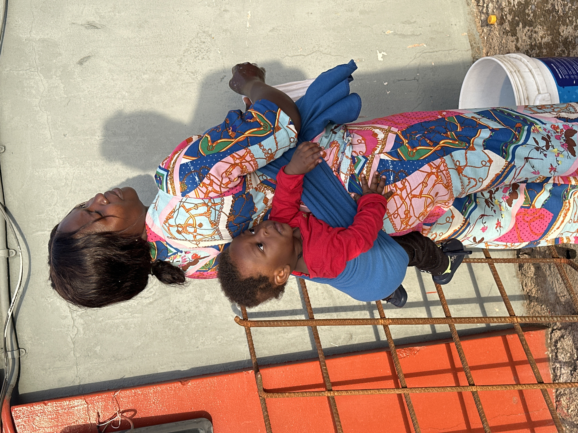 The image, set in Gura Top, Nigeria, captures an adult woman holding a young child outdoors on a bright, sunny day. The woman, who has dark skin and black hair pulled back, is seen from the chest up, looking to her left with a serene expression. She is wearing a colorful garment, likely a dress or tunic, adorned with intricate patterns in blue, pink, and gold, featuring chain links and decorative elements.
The child, a toddler with dark skin and short, curly hair, is held securely by the woman. Dressed in a red long-sleeved shirt and blue pants, the child faces away from the camera, looking upwards and to the left with a curious or engaged gaze. Their small hands rest gently on the woman's patterned clothing.
The subjects are positioned on a light gray concrete surface that shows cracks and some weathering. Strong, clear shadows on the ground indicate bright daylight and sunny weather. In the immediate foreground, a rusty metal grate with square sections is visible, suggesting a railing or a floor section of an elevated area. Behind this grate, a vibrant orange rectangular structure can be seen. To the upper right, a white bucket with a partially visible blue label rests on the concrete. A slender silver cable runs along the left side of the frame. The scene conveys a calm, everyday moment between a caregiver and a child.