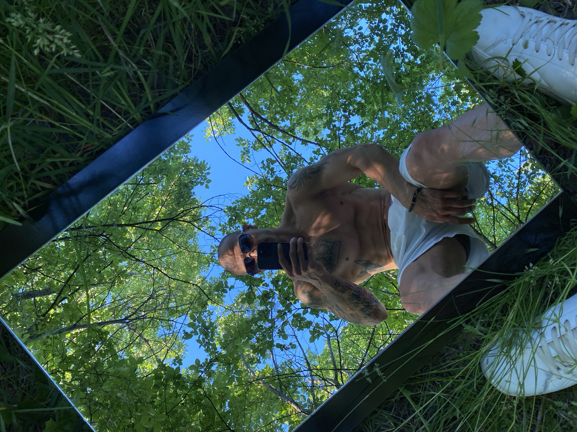 The image shows a man's reflection in a mirror placed in a grassy outdoor setting, presumably a park or wooded area. The man is shirtless and wearing sunglasses, shorts, and white shoes. He is holding a phone in his hand, presumably taking a selfie. The mirror is reflecting the sky and trees above. The location is unknown and there is nothing in the image to suggest that it is in Pleven, Bulgaria.