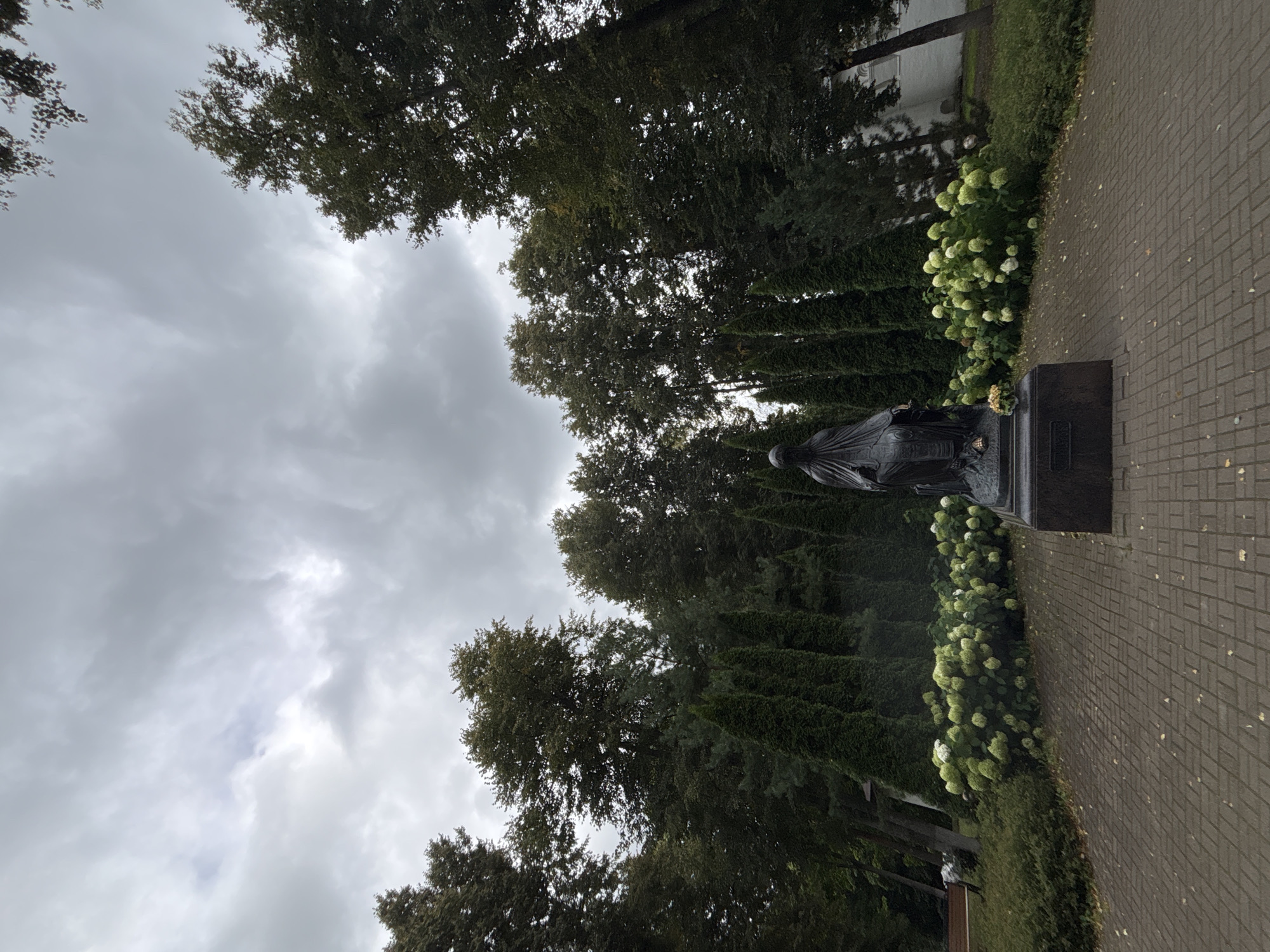 This image depicts a dark bronze statue of a seated cloaked figure, possibly a religious or historical personage, in the foreground. The statue is situated on a dark stone pedestal, which has an inscription that is illegible due to its dark color and the resolution of the image. There are a few small, light-colored flowers placed at the base of the statue.
The statue is surrounded by a semicircular arrangement of lush, white and green hydrangea bushes. Behind the hydrangeas, a line of tall, dark green cypress trees stands sentinel, creating a backdrop. Beyond the cypress trees, taller deciduous trees with dark green foliage form a dense forest, partially obscuring a white building on the far right of the frame.
The ground in the immediate vicinity of the statue and hydrangeas is paved with light brown and grey brickwork, laid in a basket-weave pattern. A few fallen leaves are scattered across the paving stones. To the left of the paved area, there is a strip of green grass, bordered by a low hedge. Further to the left, a wooden bench is visible, positioned beneath the shade of some trees.
The sky above is overcast with heavy, grey clouds, suggesting a cloudy or possibly rainy day. The overall lighting is subdued, contributing to a somber atmosphere.
The statue is located in the territory of the Savvino-Storozhevsky Monastery in Zvenigorod, Russia. This specific statue is believed to be a monument to Princess Anna Kashinskaya.
