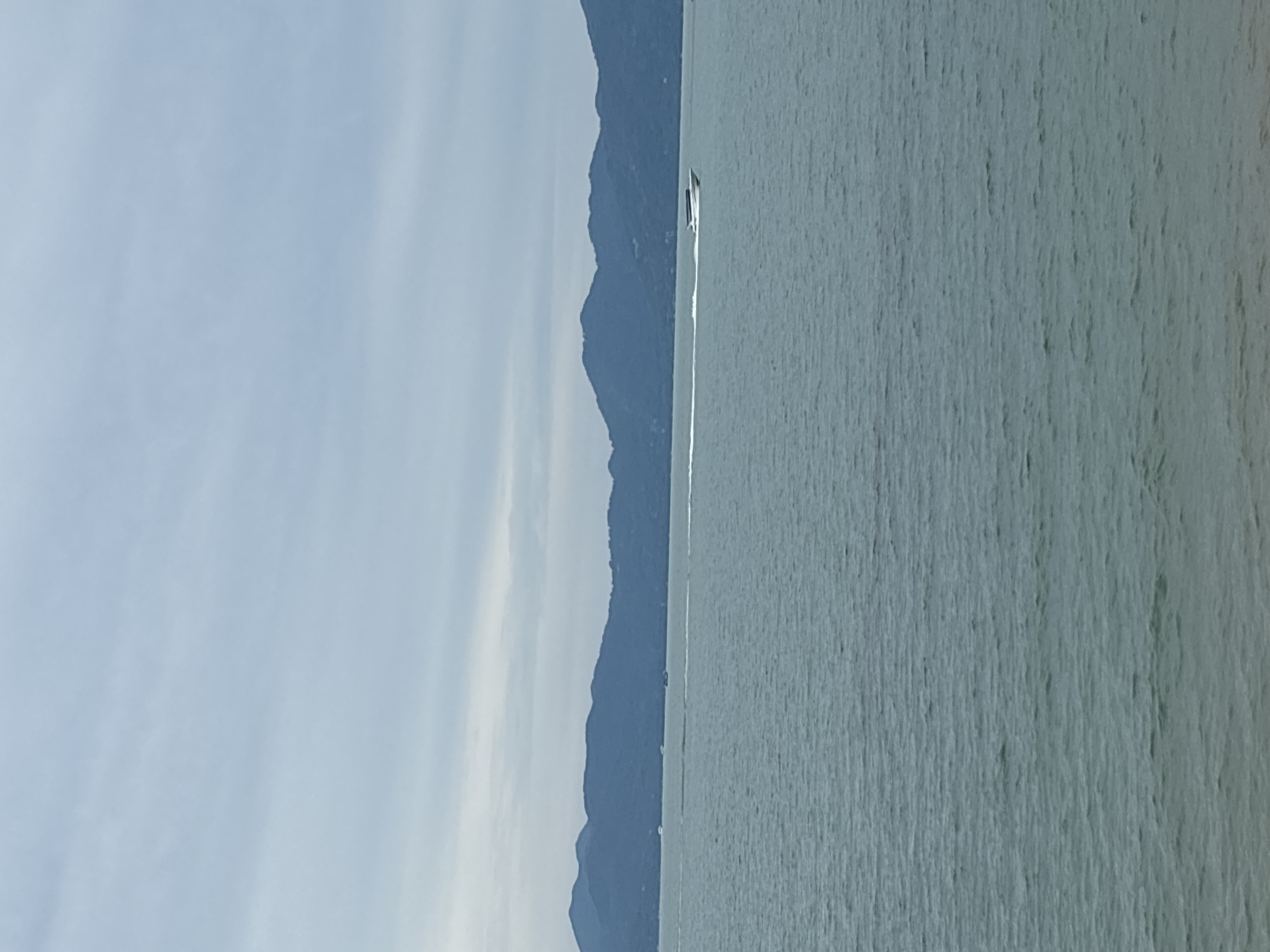 The image depicts a wide expanse of choppy, grey-green water under a cloudy sky. In the distance, a range of dark blue, rounded mountains forms the horizon. A white motorboat is visible in the middle ground, moving from left to right across the frame, leaving a distinct white wake behind it. The boat appears to be a speed boat with a dark canopy. The foreground shows a faint hint of a sandy or rocky shore. The overall atmosphere is overcast and still. This scene is likely a coastal view in Baan Bo Phut, Thailand, given the presence of the sea, islands, and type of boat commonly seen in tourist areas.