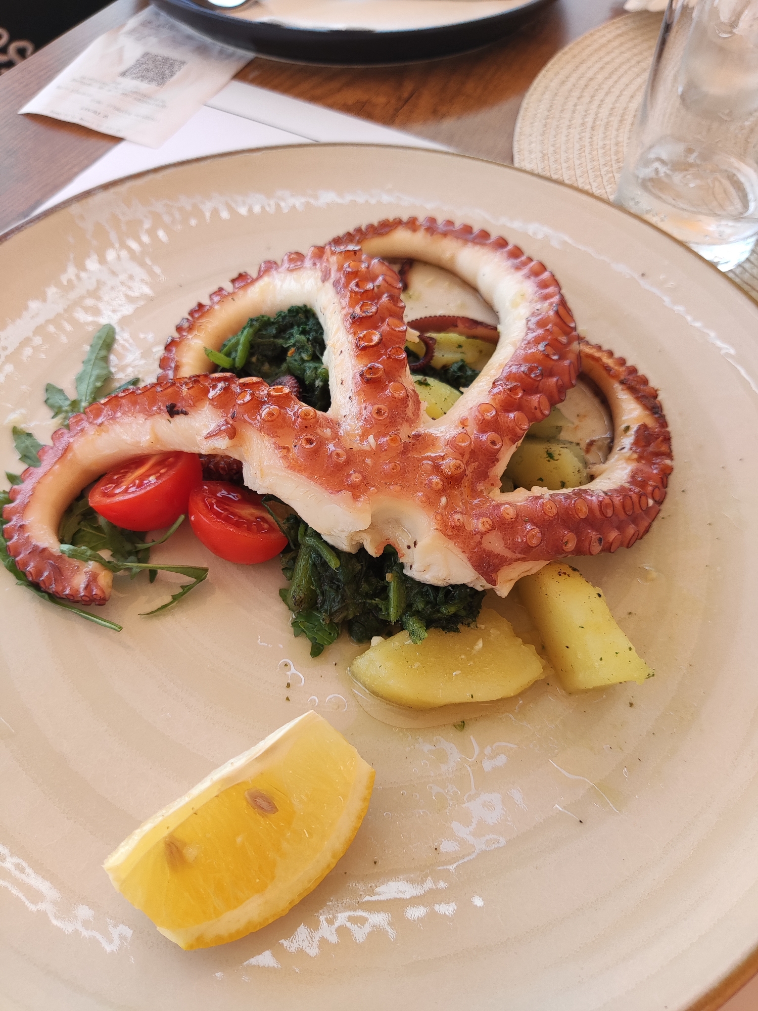 A close-up overhead shot shows a serving of grilled octopus tentacles served on a creamy beige plate. The octopus tentacles are arranged in a circular fashion, with the suckered undersides prominently displayed. They are a reddish-brown hue, indicating they have been grilled. Nestled beneath the octopus is a bed of dark green, wilted greens, possibly spinach or kale, interspersed with chopped herbs. Several halved cherry tomatoes, red and ripe, are scattered among the greens. To the right and bottom of the octopus are chunks of boiled or roasted potatoes, golden yellow with flecks of green herbs. A single, bright yellow lemon wedge sits in the lower left foreground, its juicy interior and a couple of seeds visible. The plate is set on a wooden table, and in the background, a partially visible receipt with a QR code and a glass of water with ice can be seen. The overall impression is a well-presented, savory seafood dish. The location is stated as Dobra Voda, Montenegro.