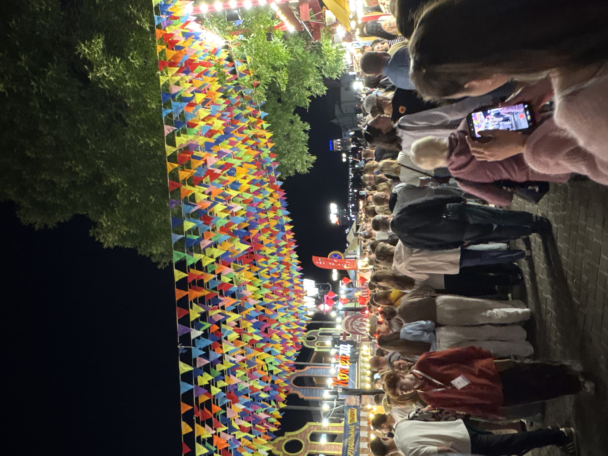 This is a medium shot, taken at night, of a crowded outdoor event, likely a festival or fair, in the city of Vitsebsk, Belarus. The scene is illuminated by string lights and the colorful flags that hang overhead, creating a festive atmosphere. In the foreground, a young woman is taking a picture with her phone of the crowd and the decorated street. She is wearing a pink fuzzy sweater. The background is filled with a diverse crowd of people, with many individuals blurred due to motion. Among the crowd, some are facing forward, while others are looking at their phones or interacting with each other. The environment includes stalls or booths with signs in Cyrillic, suggesting a local setting. The overall impression is a lively and bustling public gathering.