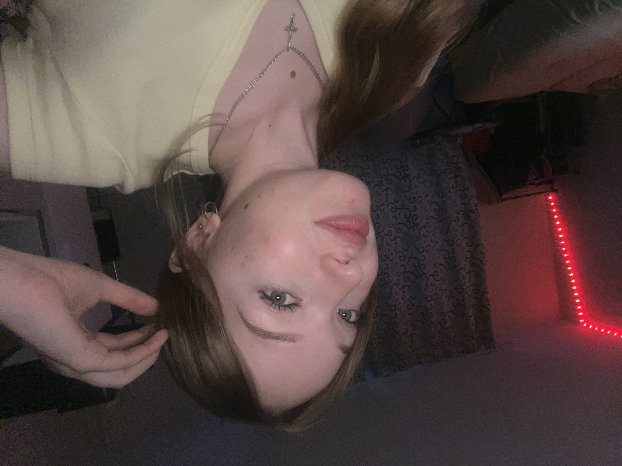 A photograph of a light-skinned female wearing a yellow V-neck shirt and a silver chain with a cross charm, situated indoors. Her face is turned towards the camera; her hair is partially obscuring her face. Her hand is partially visible near her hair. The background includes a patterned curtain, a black object with visible linear features, and a red LED strip. There is no verifiable information to associate this image with the city of Chaikovsky, Russia.