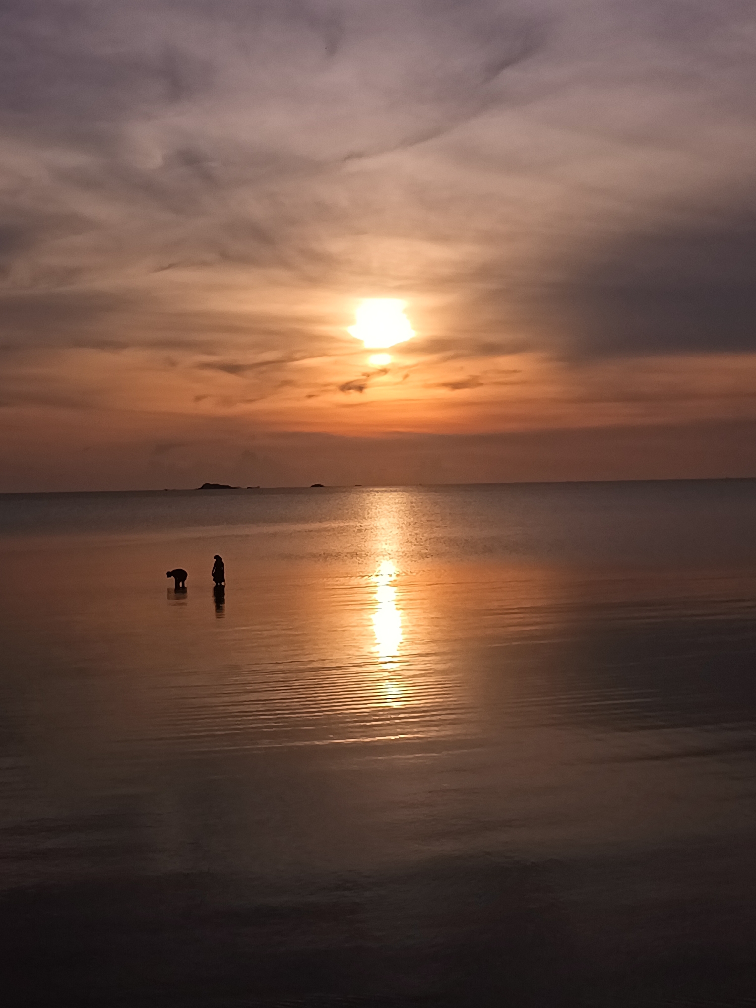 The image captures a sunset scene in Baan Wok Tum, Thailand. Two figures are silhouetted against the golden water, one bending over and the other standing upright. The sea reflects the sunlight, creating a bright path from the sun to the foreground. A small landmass is visible on the horizon. The sky is filled with clouds tinged with orange and gray.
