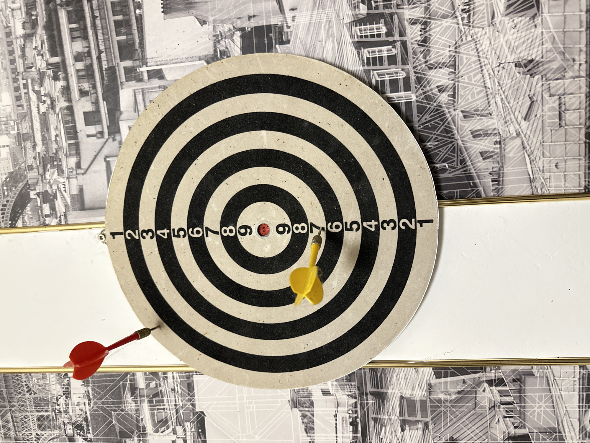 An overhead, slightly angled shot shows a dartboard mounted on a white surface, with a black and white cityscape as the background. The dartboard is circular, with alternating black and cream-colored concentric rings. Numbers from 1 to 9 are printed in black along the right side of the dartboard. Two darts are visible. A red dart with two fletchings is stuck in the upper left portion of the dartboard, slightly above the number 2. A yellow dart with three fletchings is stuck in the lower left portion of the dartboard, between the numbers 7 and 8. A small red circular bulls-eye is visible in the center of the dartboard. The background is a black and white photograph of a dense urban environment with many buildings and some sort of arched structure, possibly the Eiffel Tower, visible in the upper left corner. There is a vertical gold frame on the left side of the image, running from top to bottom, partially obscuring the dartboard and background. There are no people or animals in the image.