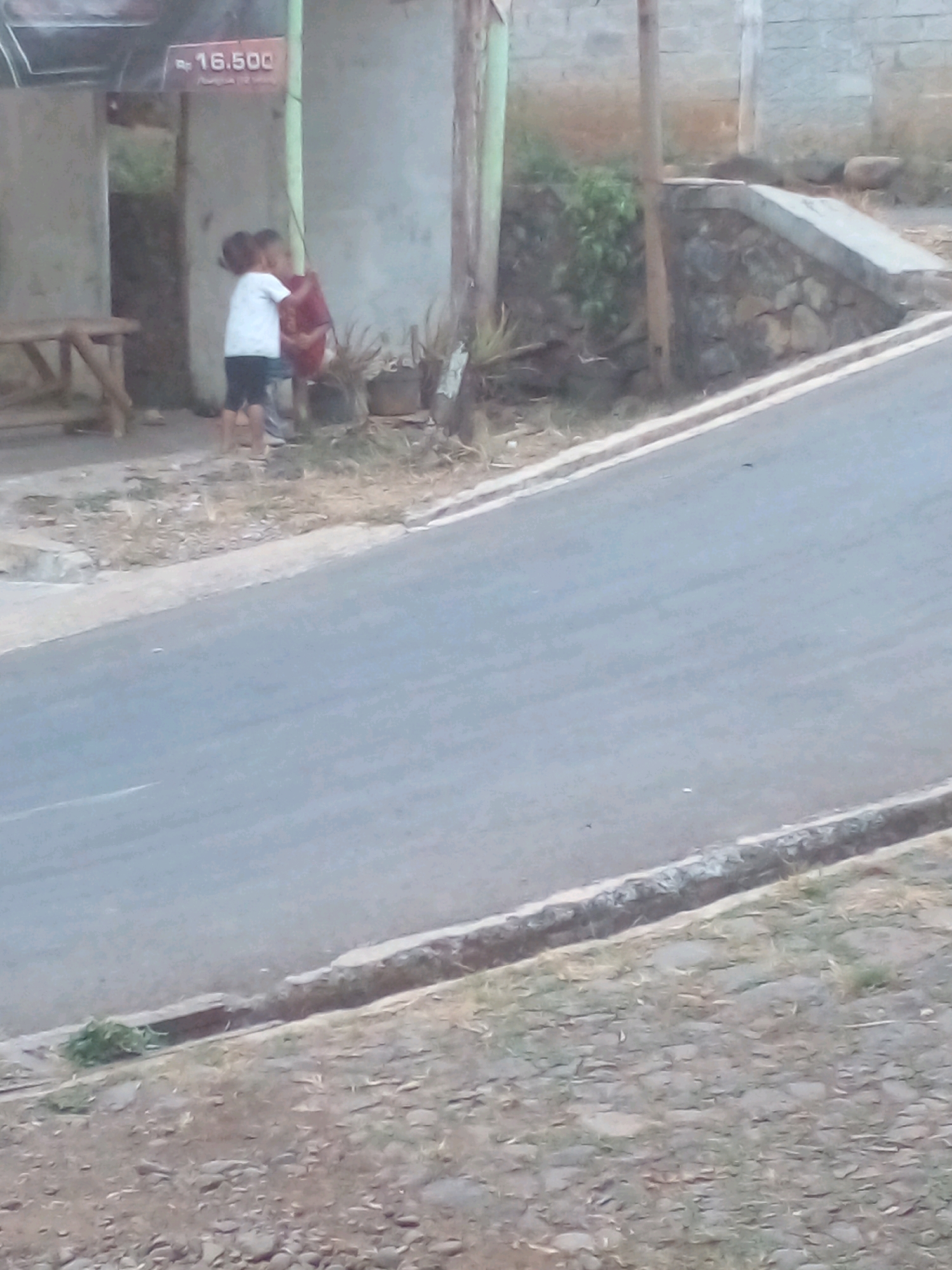 In Pungangan, Indonesia, two young children stand near a weathered concrete structure and a wooden bench. One child, wearing a white t-shirt and dark shorts, embraces another child who is holding a red object, possibly a toy or a bag. Behind them, a green pole supports a sign displaying "Rp 16.500". The ground is covered in dry grass and dirt, with a grey asphalt road bordering the scene. A stone wall and vegetation are visible in the background.