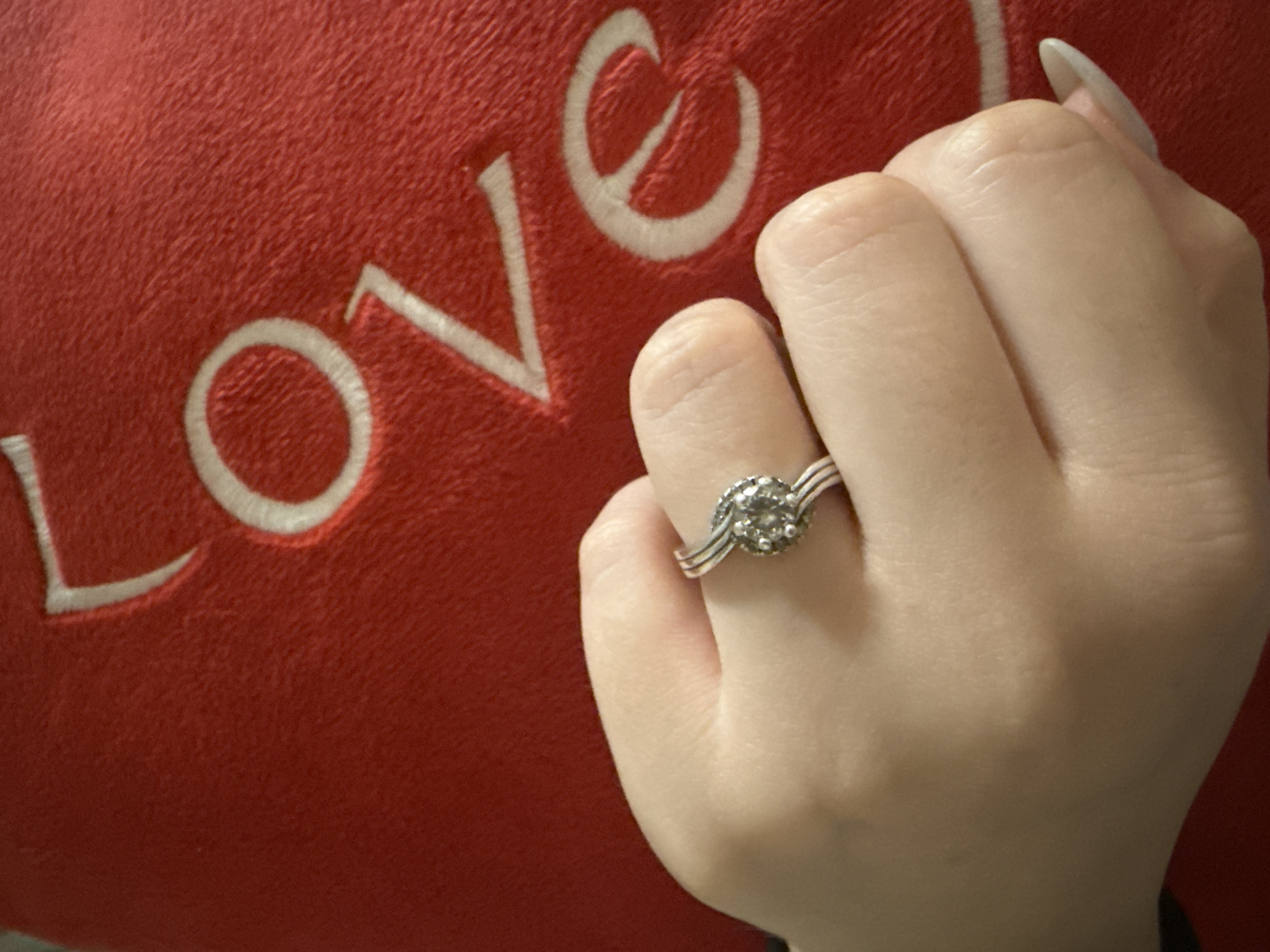 A close-up shot shows a hand wearing a diamond ring, held against a red pillow with the word "LOVE" embroidered on it in white.