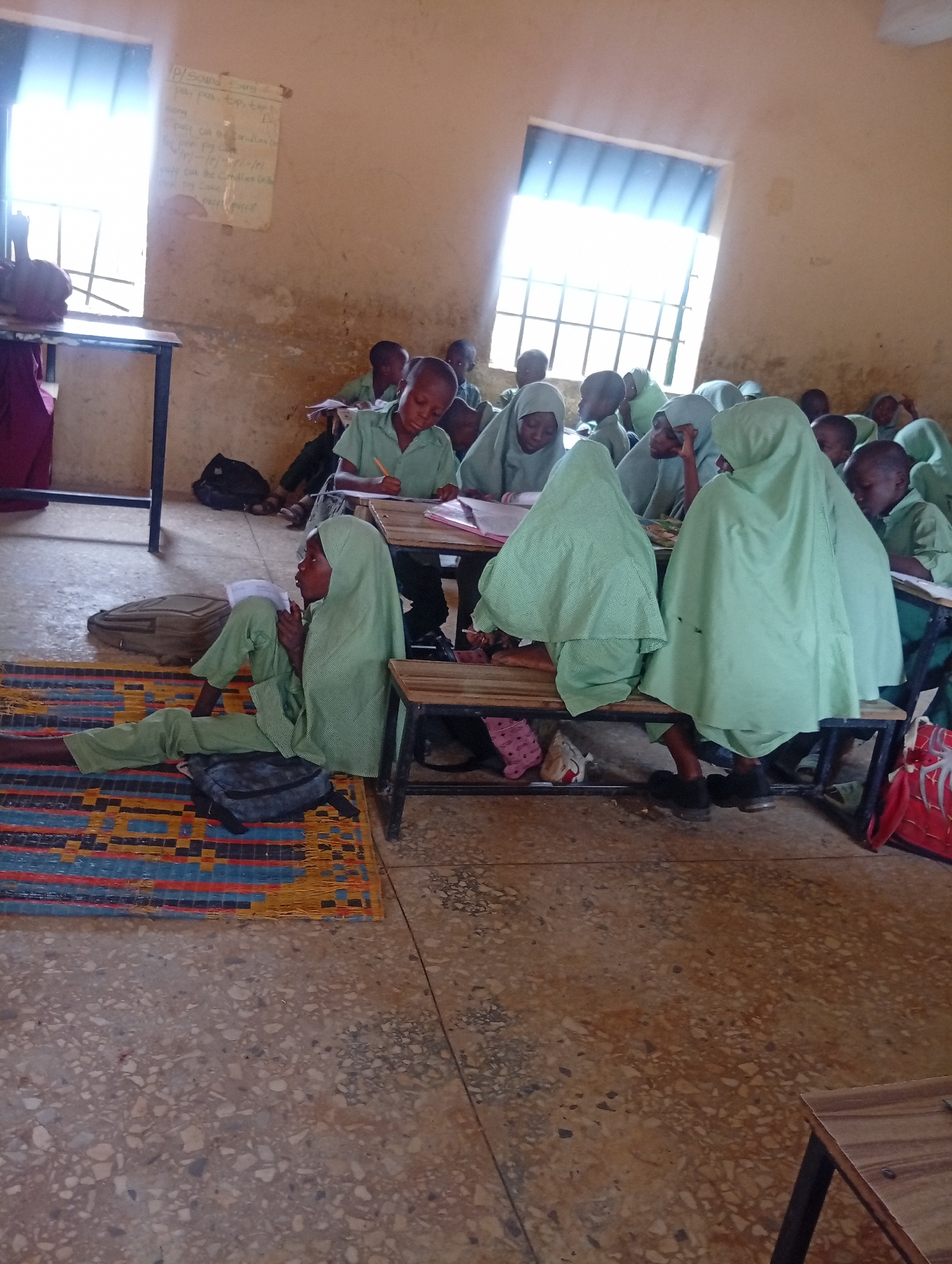 The image depicts a classroom scene in a school located in Dutse, Nigeria, likely an educational facility catering to young girls. Several female students, predominantly wearing light green hijabs and uniforms, are seated at desks and on the floor, engaged in learning activities. Some are writing or reading from books, while others appear to be listening or observing. The classroom environment is basic, with bare walls and a concrete floor. Natural light streams in from windows covered by slatted blinds. Educational materials, such as a whiteboard with writing, are visible on the walls. Scattered on the floor are personal belongings like bags and mats, suggesting a practical and perhaps resource-limited setting. The overall impression is one of diligent study within a humble educational space.