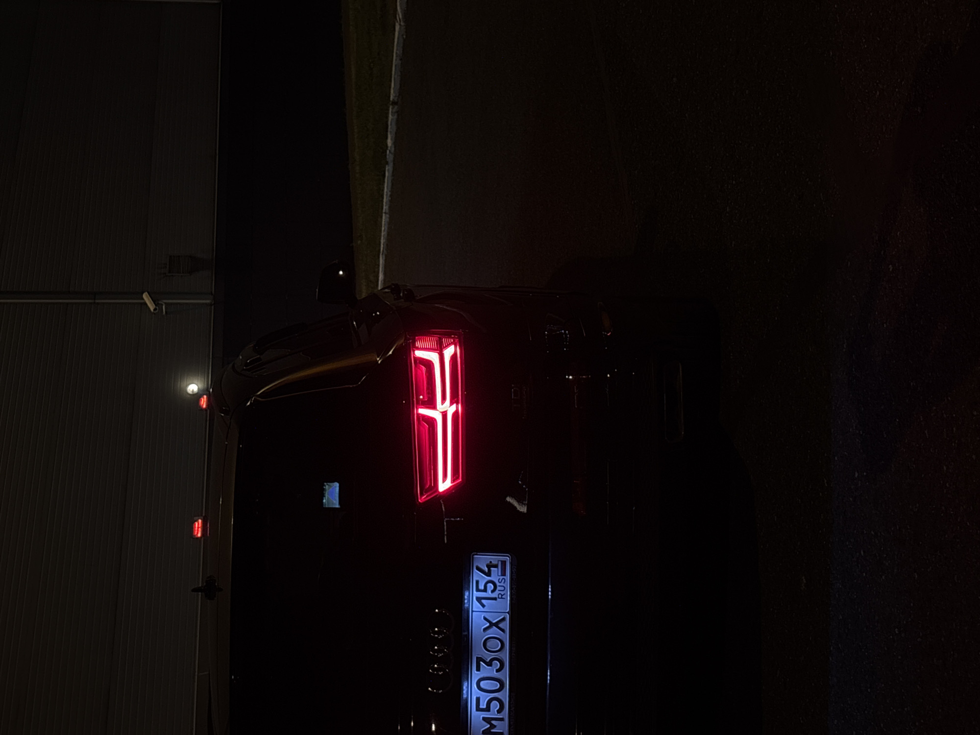 The image shows the rear of a dark-colored Audi Q7 parked at night on a paved surface, possibly a parking lot, adjacent to a grassy area and a building with a large corrugated metal facade. The car's taillights are illuminated with a distinct red LED pattern. A license plate is visible on the rear of the vehicle, displaying the alphanumeric characters "M503OX 154" with "RUS" below it. The scene is dimly lit, with the primary light sources being the car's taillights and some ambient artificial lighting from the building. There are no people visible in the image. The overall environment appears to be an industrial or commercial area in Novokuznetsk, Russia, given the presence of the building and the Russian license plate.