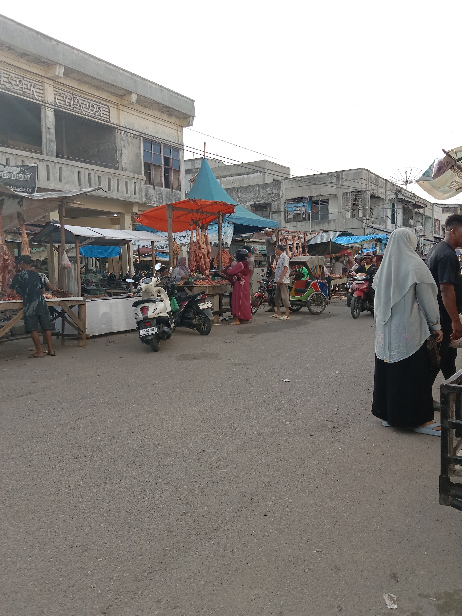 The image depicts an open-air market scene in Lhokseumawe, Indonesia. Several stalls are set up along a street, selling meat, with carcasses hanging from orange and blue tarps. People are shopping, some wearing traditional Muslim attire, including a woman in a grey hijab and black clothing in the foreground. Several motorbikes and a colorful tricycle are parked or moving through the scene. The buildings in the background appear old and somewhat dilapidated, with some having empty window spaces. The sky is overcast.
