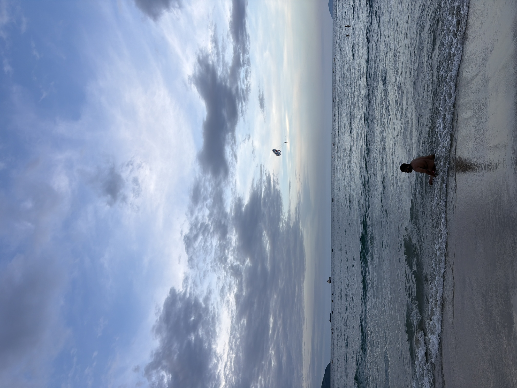 In Đà Nẵng, Vietnam, this image captures a serene beach scene at dusk. The sky is a canvas of soft blues and grays, with dramatic clouds stretching across the horizon. The sea is calm, with gentle waves lapping at the sandy shore.
In the foreground, a lone man sits on the beach, partially submerged in the shallow water, facing away from the camera. His posture suggests relaxation or contemplation as he gazes out at the sea. In the middle distance, a parasailer is visible in the sky, a splash of color against the muted tones of the sky, indicating recreational activity. Further out at sea, several boats are scattered, some appearing to be fishing vessels, while a larger ship is visible on the horizon, suggesting maritime activity. The overall atmosphere is tranquil, hinting at the end of a day at the beach.