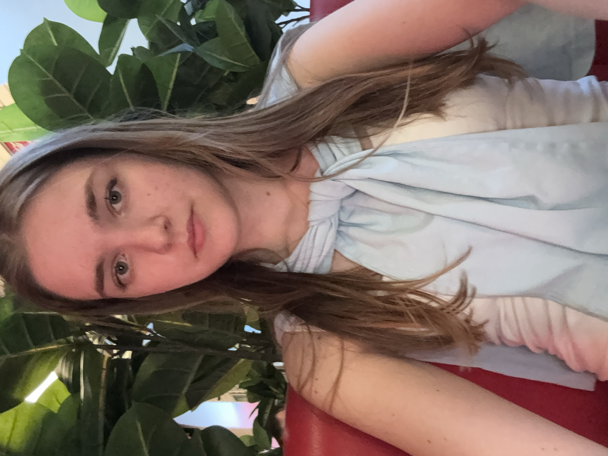 A young woman with light brown hair, fair skin, and light-colored eyes is the primary subject. She is wearing a light blue garment. Large green leafy plants are visible in the background. She is seated on a red surface. The location in the city of Perm, Russia is undeterminable from the image.