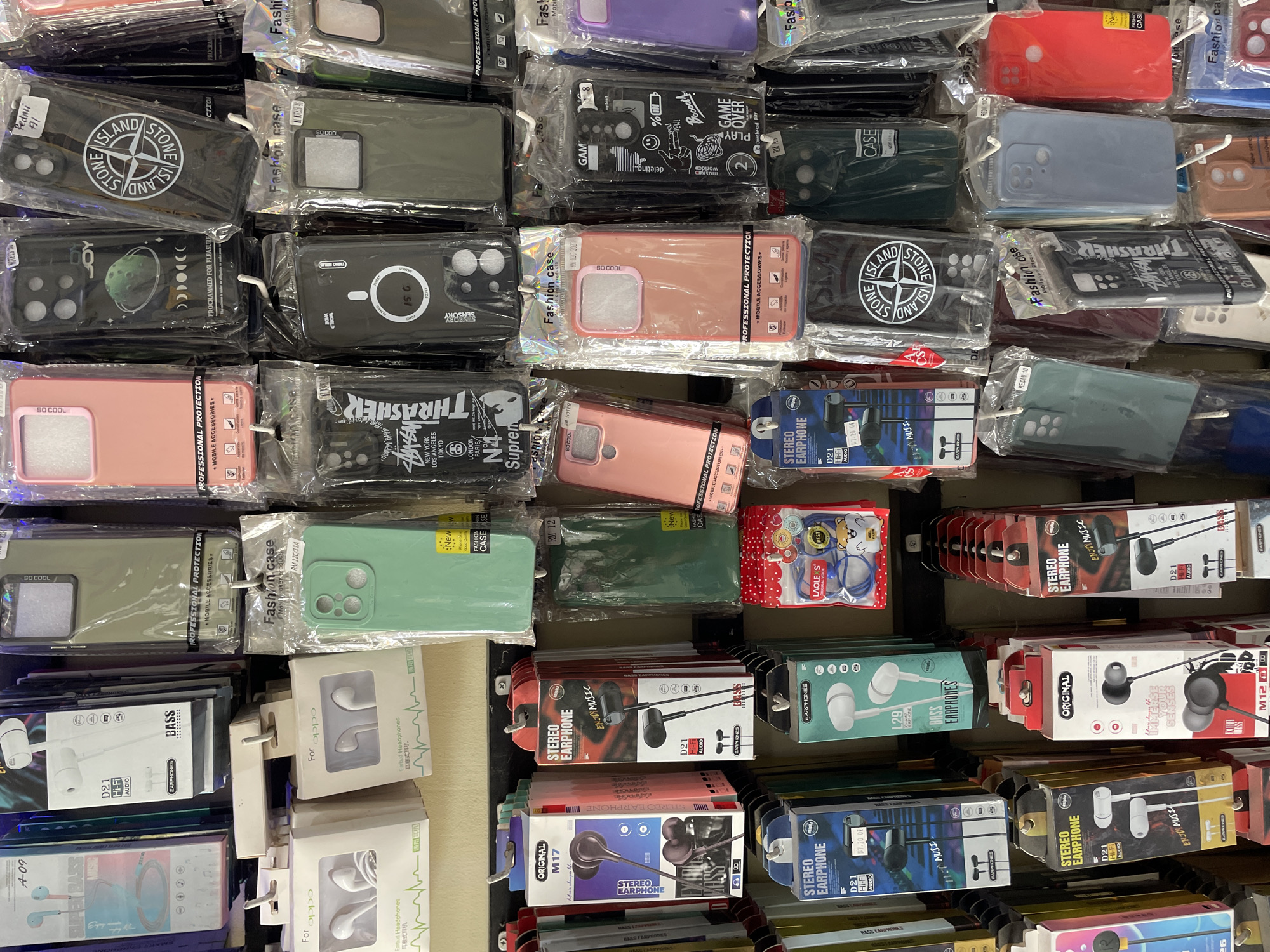 The image shows a display of mobile phone accessories in a shop, likely in Pasar Paloh, Indonesia. The display features rows of phone cases and earphones packaged in clear plastic and cardboard boxes. The phone cases are varied in color and design, with some featuring branded logos and graphics. The earphones are also packaged with brand names and specifications like "D21 Hi-Fi Audio" and "Bass Earphones". The overall environment suggests a retail setting where these items are being sold, with the products hung from hooks on a wall. The lighting is bright and evenly distributed, illuminating the colorful packaging and diverse range of products.