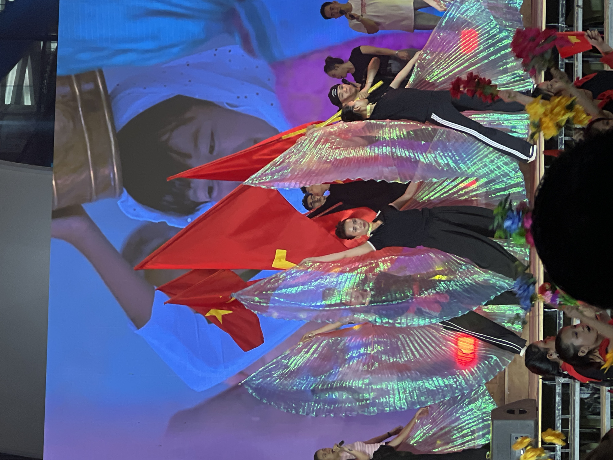 This media file captures an indoor stage performance or cultural event, likely taking place in the evening. The scene is dominated by a large projection screen in the background, which displays a close-up image of a child or young person with a reflective surface.
On the stage, numerous performers are visible, ranging from young adults to possibly younger children on the far right. Many of the performers are dressed in black attire, including black shirts and pants, with some pants featuring white stripes along the sides.
The key props in the performance are large, red flags adorned with a yellow star, unequivocally identifying them as the national flags of Vietnam. Several performers are holding and waving these flags. Additionally, many performers are holding large, shimmering, iridescent fan-like objects. These props reflect various colors from the stage lighting, creating a vibrant effect that includes hues of green, blue, and pink.
On the upper right side of the image, one male individual is seen holding a microphone, suggesting he might be an announcer, host, or a singer in the performance. To his left, another person also appears to be participating in a similar role. On the far right, some performers are wearing colorful floral headpieces or holding bouquets of artificial flowers, adding to the festive atmosphere. A speaker box is also visible on the stage floor.
The overall impression is one of a lively and patriotic celebration, with the Vietnamese flags and coordinated performance signifying a cultural event. The colorful lighting and theatrical setup suggest a well-organized show.
