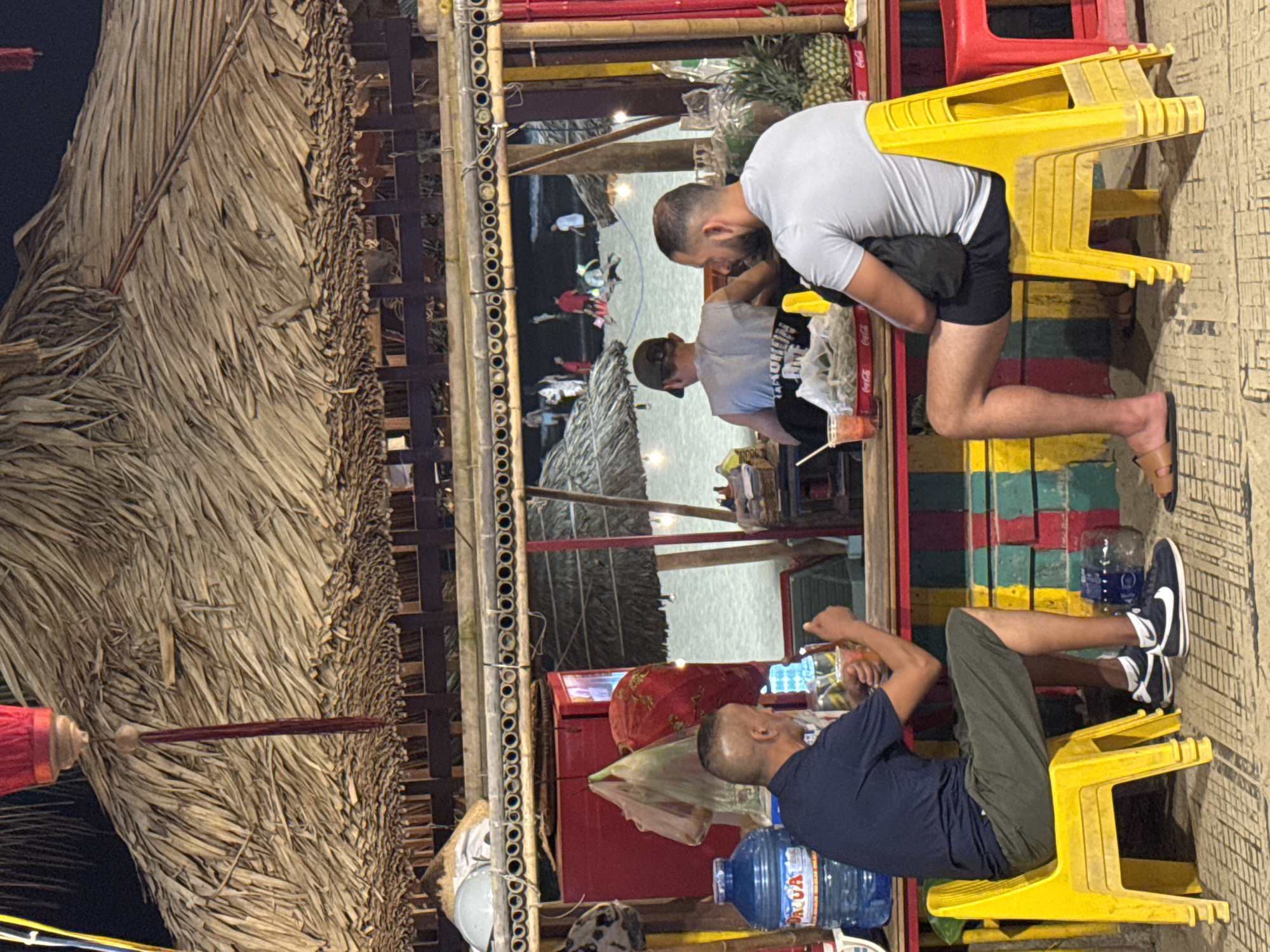 The image captures an informal outdoor scene in Đà Nẵng, Vietnam, possibly at a seaside establishment. Two men are seated on stacked yellow plastic stools, with one of them looking at a mobile phone. Another man stands nearby, also engaged in a conversation. The setting appears to be a bamboo structure with a thatched roof, reflecting the local architectural style. In the background, a beach with other people is visible, creating a relaxed, touristy atmosphere. There are beverage supplies, water bottles, Coca-cola and pineapples on the countertop.