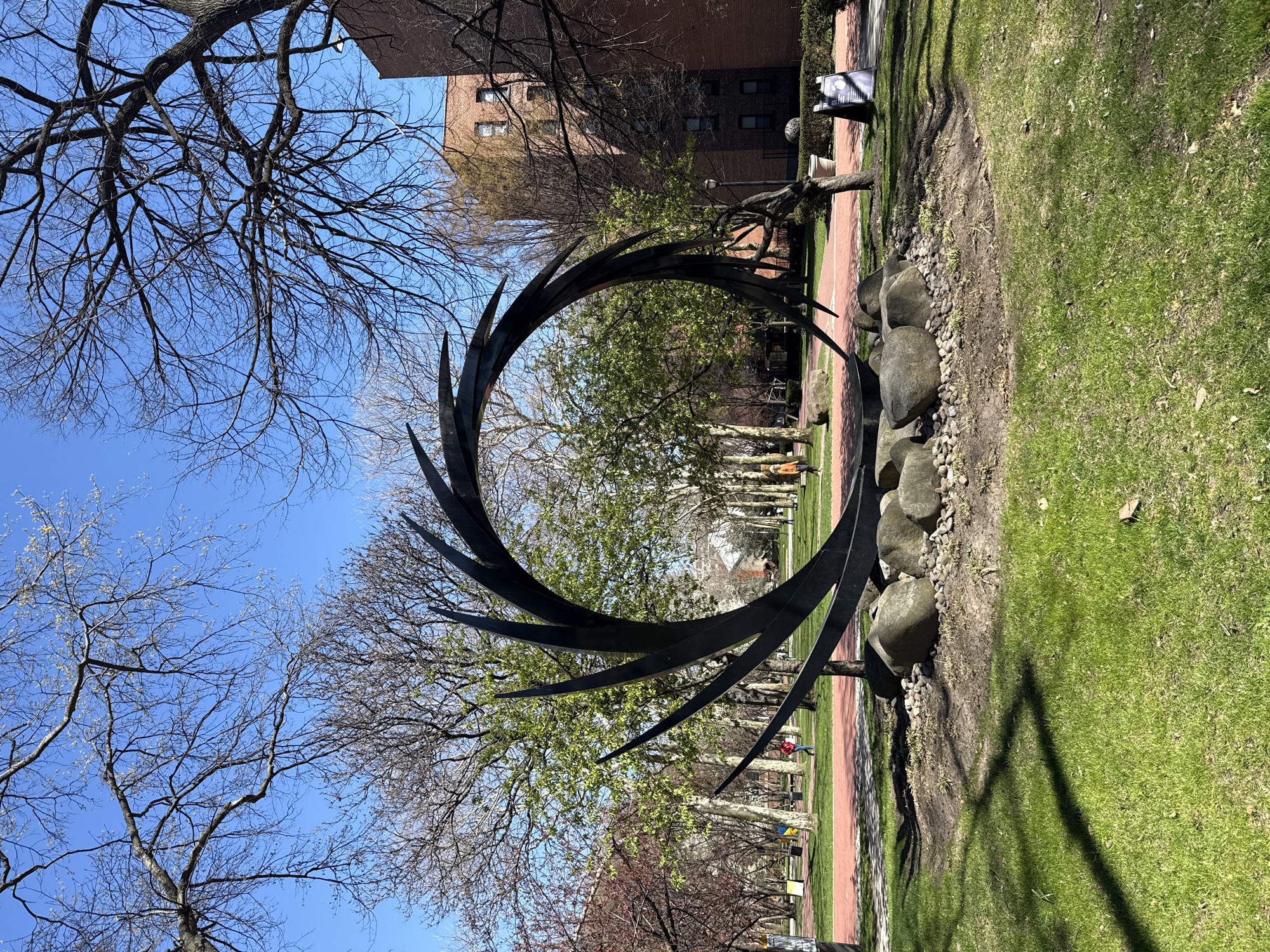 The image depicts a sculpture in an outdoor setting. The sculpture is a large, circular, metal structure with sharp, blade-like protrusions extending from its circumference. The sculpture is surrounded by a line of rocks. The background consists of trees, grass, a brick pathway, and buildings. People are walking in the background. The location is identifiable as a park or garden in the city of New York, United States of America.