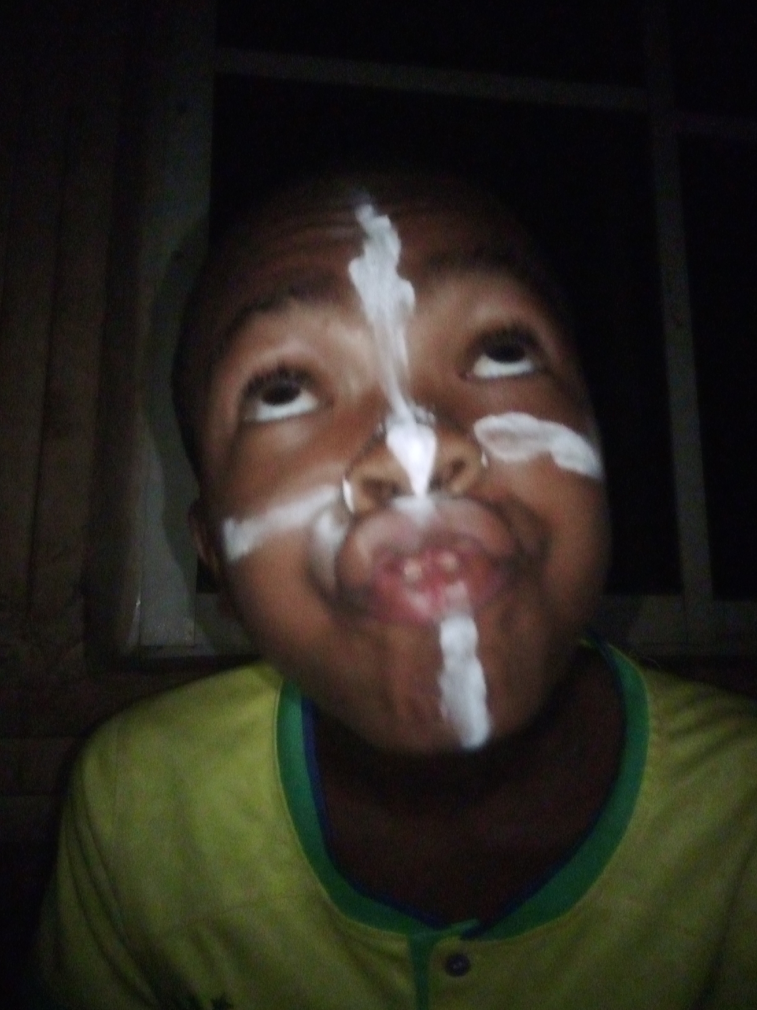 A medium close-up portrait features a young person, likely a child or early adolescent, with dark skin, gazing upwards with a playful, curious, or slightly mischievous expression. Their mouth is partially open, revealing a few teeth. The face is adorned with white markings: a prominent vertical stripe extends from the forehead, across the bridge of the nose, and down to the upper lip, intersected by two shorter horizontal stripes across each cheek, forming a cross-like design. A small, metallic-looking ring is visible through the septum of the nose. The individual wears a light-colored (appearing yellow or lime green) short-sleeved shirt with a green and blue collar trim.
The background is predominantly dark and indistinct, suggesting an indoor environment or nighttime. A window frame or similar dark rectangular structure is partially visible in the upper right. The scene is dimly lit, with the light source seemingly illuminating the front of the person's face. The face paint implies participation in a celebration, cultural event, or imaginative play, potentially within the context of Onitsha, Nigeria. No visible text is present in the image.