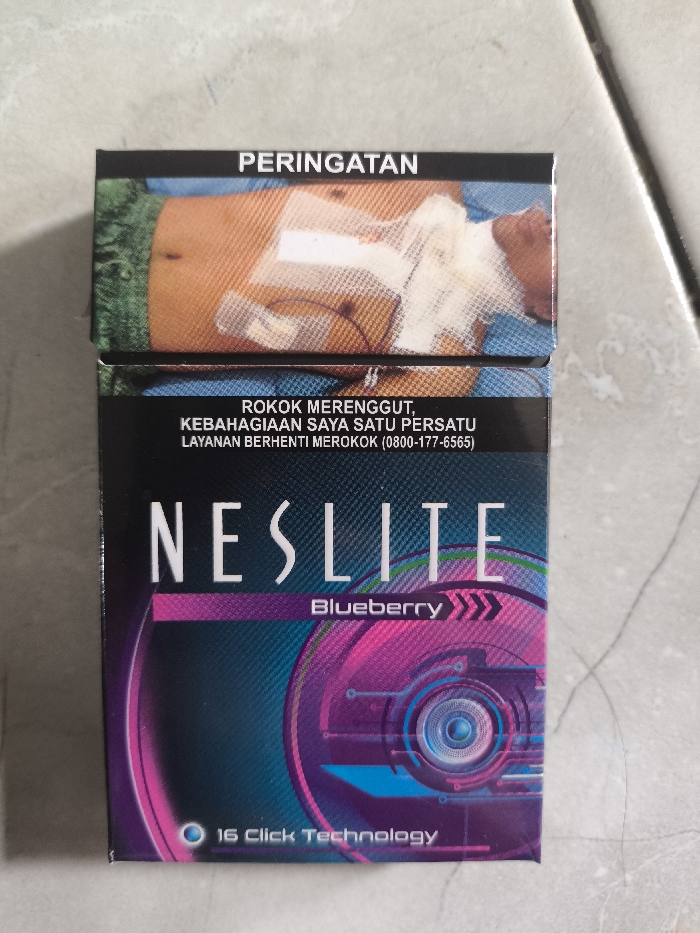 A pack of "NESLITE Blueberry" cigarettes sits on a tiled surface. The package features a futuristic design with purple and teal accents. Below the brand name, "Blueberry" is printed with arrow motifs. At the bottom, "16 Click Technology" is visible. The top of the pack has a graphic health warning: an image of a person in a hospital setting with bandages and tubes. Above the image is the word "PERINGATAN," and below is a line of text that reads "ROKOK MERENGGUT, KEBAHAGIAAN SAYA SATU PERSATU LAYANAN BERHENTI MEROKOK (0800-177-6565)".
