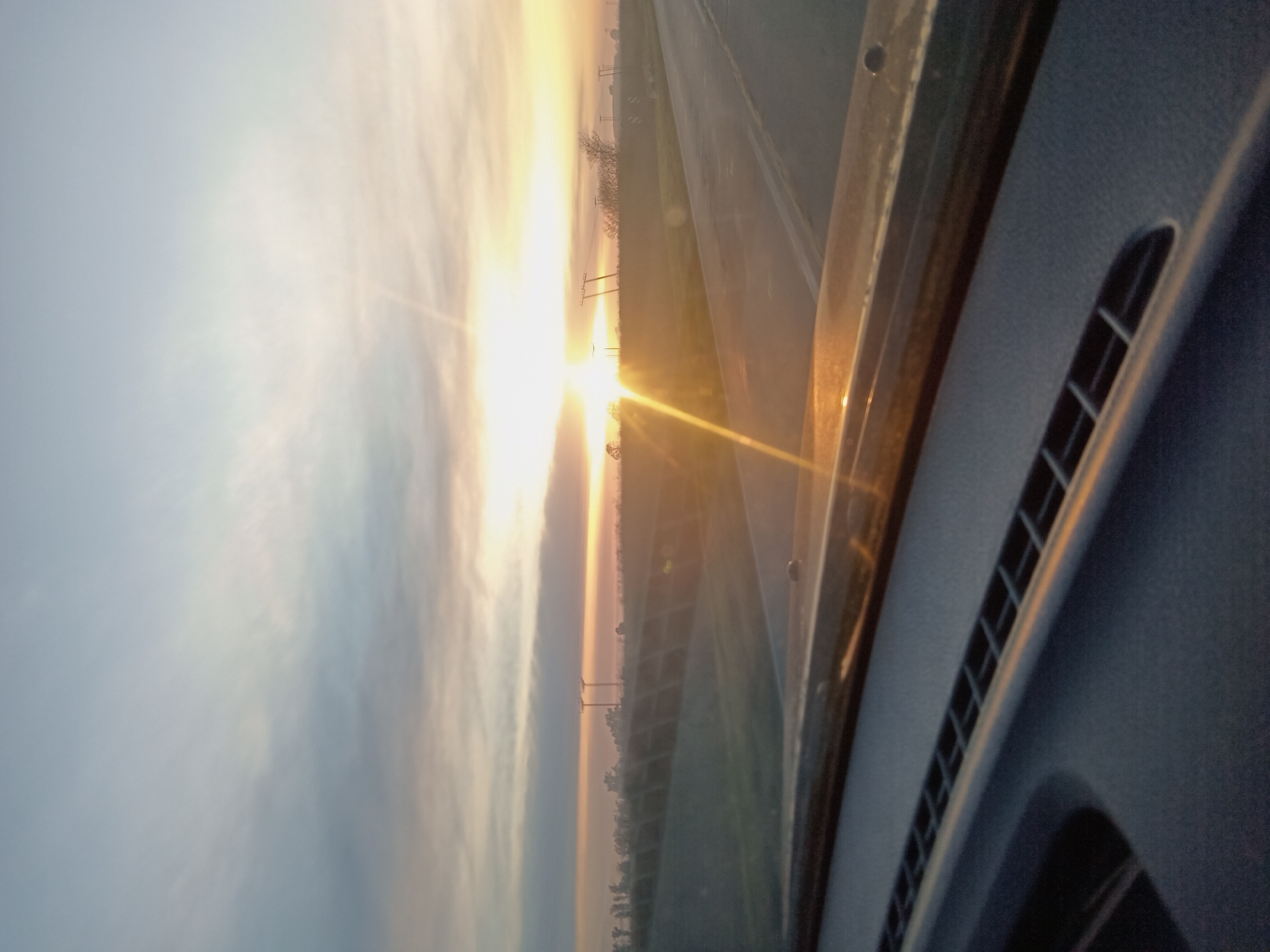 An exterior, medium shot captures a view from inside a vehicle looking out at a road and the sky at sunset. The foreground is dominated by the dark dashboard of the car, with a textured surface and vents. Beyond the dashboard, a paved road curves to the right, with a grassy verge and a fence on the left. The horizon is marked by a line of trees and utility poles. The sky is a soft gradient of blues and oranges, with the sun low on the horizon, casting a bright glare that creates lens flare. The overall scene evokes a sense of travel and the end of the day.