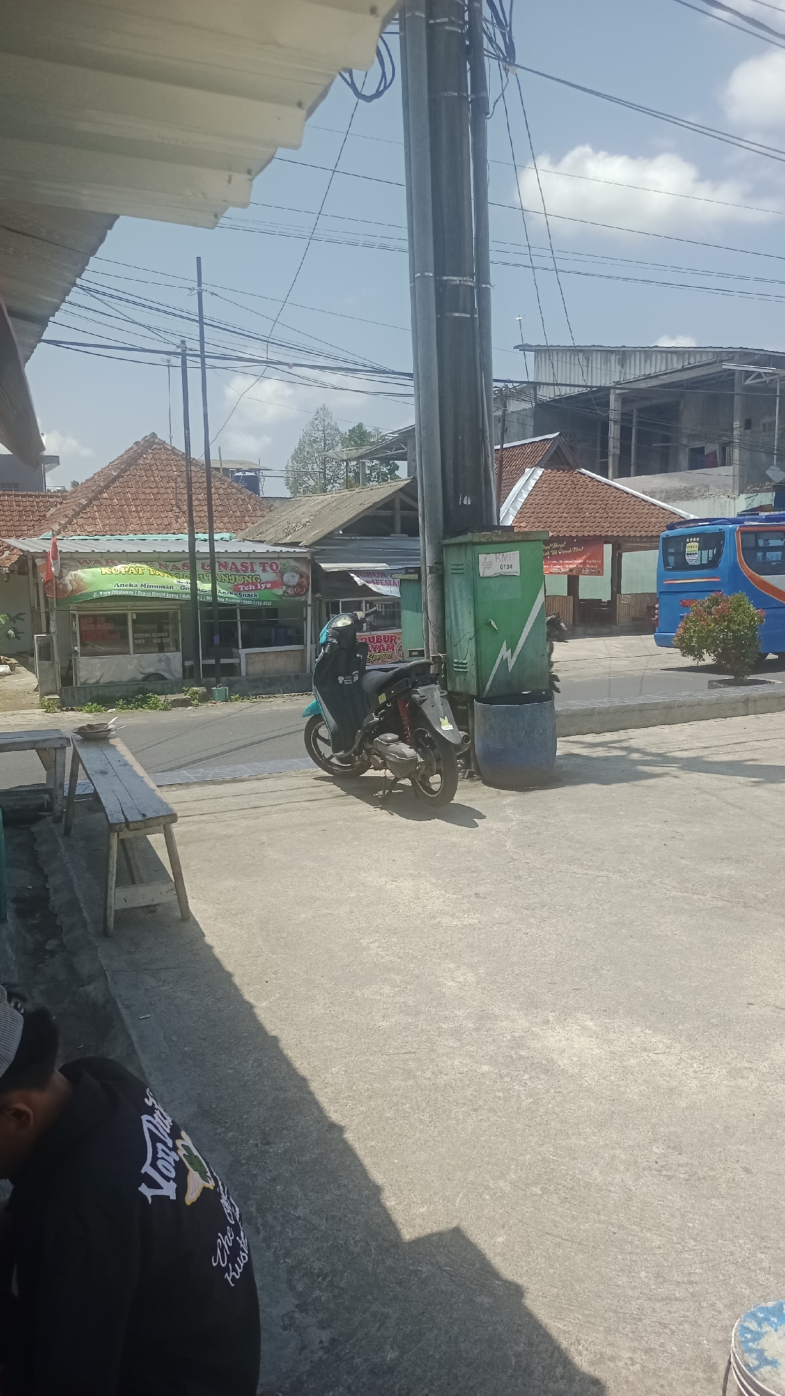 The image depicts a street scene in Pakemitan, Indonesia. In the foreground, a wooden bench is situated on a concrete surface. A scooter is parked to the right of the bench, facing the street. Behind the scooter, a tall utility pole with numerous cables is prominent, alongside a green electrical box and a blue cylindrical trash receptacle. To the left of the utility pole, a building with a red-tiled roof appears to be a food establishment, with signage visible. Across the street, more buildings with red-tiled roofs are present. In the background, a blue bus with orange and white accents is parked. The sky is bright blue with scattered clouds. A person wearing a black hooded sweatshirt with white lettering is partially visible in the lower left corner of the image.