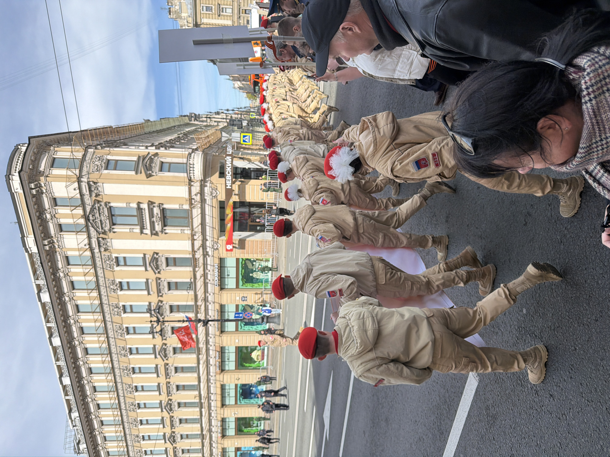 A street view in Saint Petersburg, Russia, shows a line of uniformed figures doing push-ups on the pavement. Each figure wears a tan uniform and a red cap with some having white pom poms. Behind them, a crowd of onlookers watches. A building with ornate architectural details runs along the left side of the frame. The street has traffic lines marked, and flags hang from the building. Pedestrians walk behind the performing group. Overhead cables cross the street.