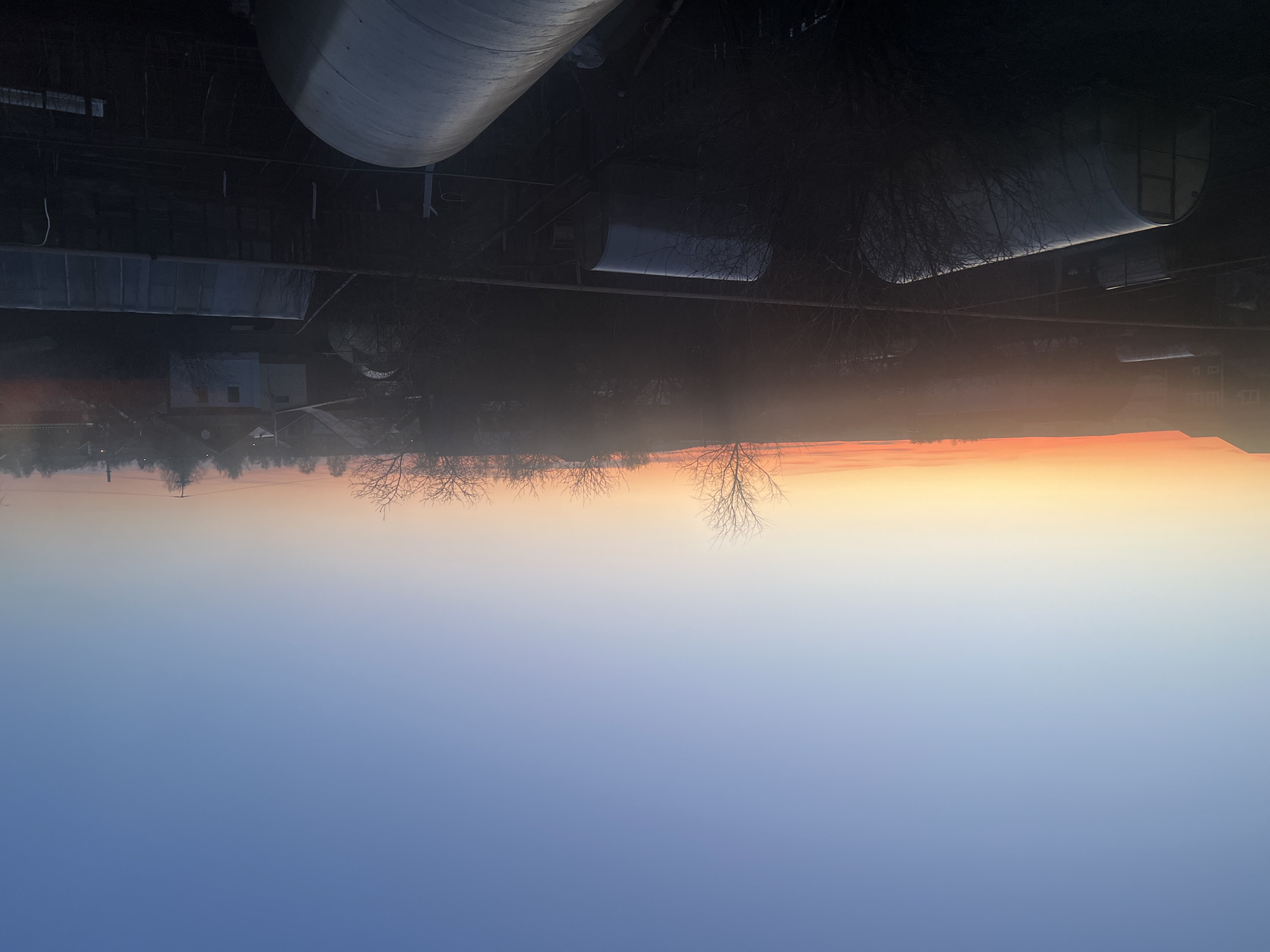 The image depicts a sunrise over the city of Votkinsk, Russia. The sky transitions from deep blue at the bottom to orange and pink at the horizon. Silhouetted trees line the horizon. Above the trees, there are industrial structures, including a large cylindrical tank and various buildings. The overall scene conveys a sense of urban life juxtaposed with a natural sunrise.