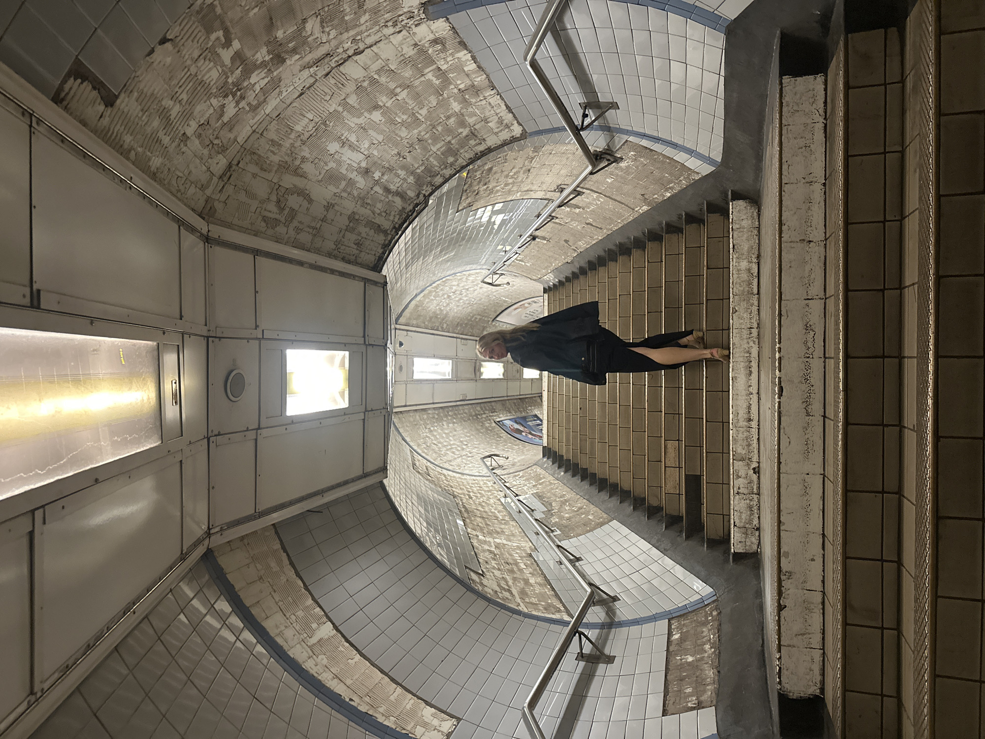 The image depicts the Aldwych tube station in London. The environment is an underground tunnel covered with white and gray tiles. A woman wearing a black coat is walking down a set of stairs. The ceiling is arched and shows signs of age, with crumbling sections. A metal handrail is visible alongside the stairs. The lighting is provided by fluorescent lights visible through openings.