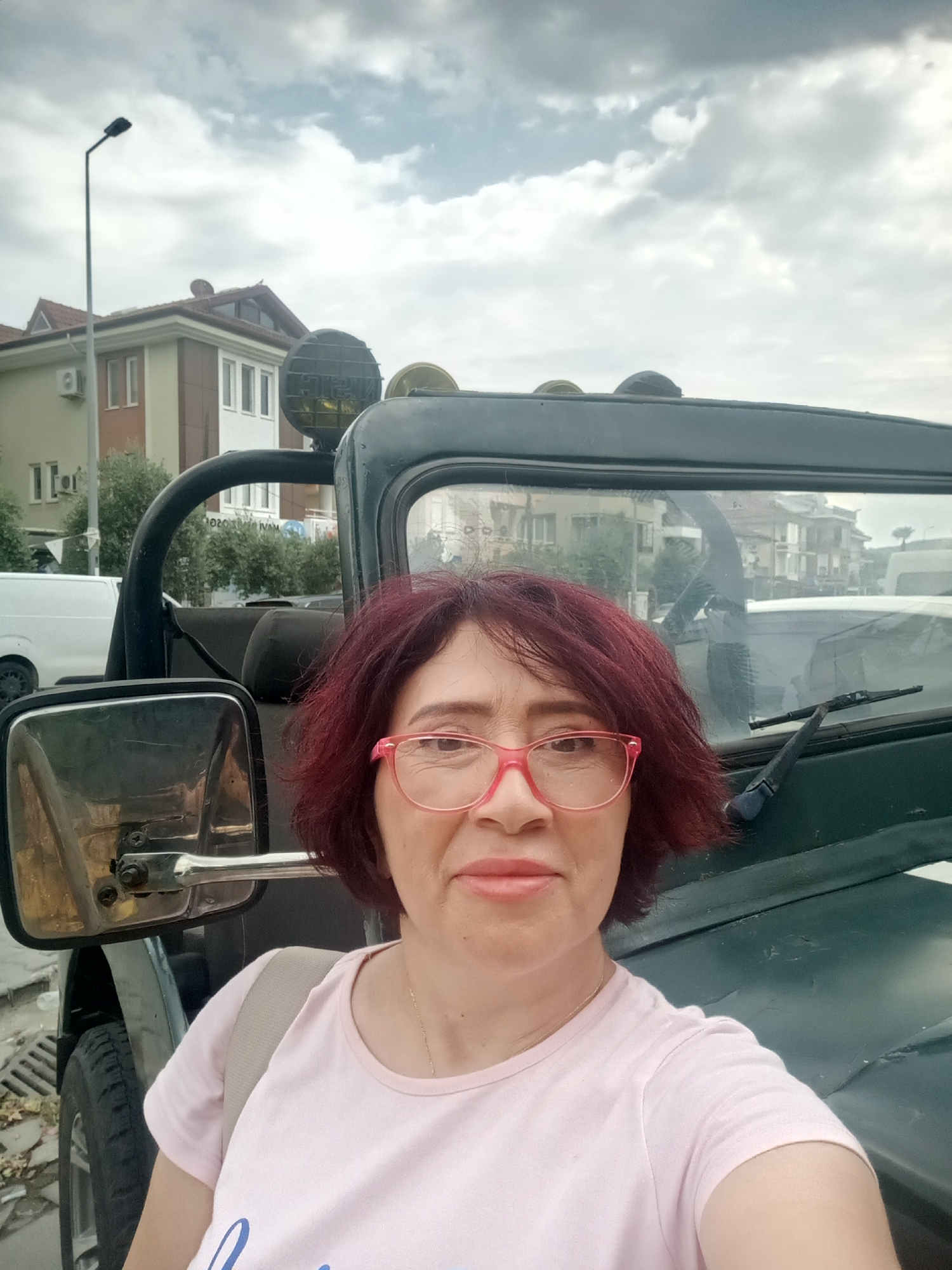 A woman with reddish-brown hair and pink-rimmed glasses is taking a selfie in front of a dark green, open-top vehicle, possibly a Jeep, parked on a street in Fethiye, Turkey. The woman is wearing a light pink t-shirt and a beige backpack. In the background, there are multi-story buildings with red tiled roofs, trees, a street lamp, and a cloudy sky. The vehicle has a roll cage, large headlights mounted on top, and a side mirror. The street appears to be paved with concrete and has a drainage grate.
