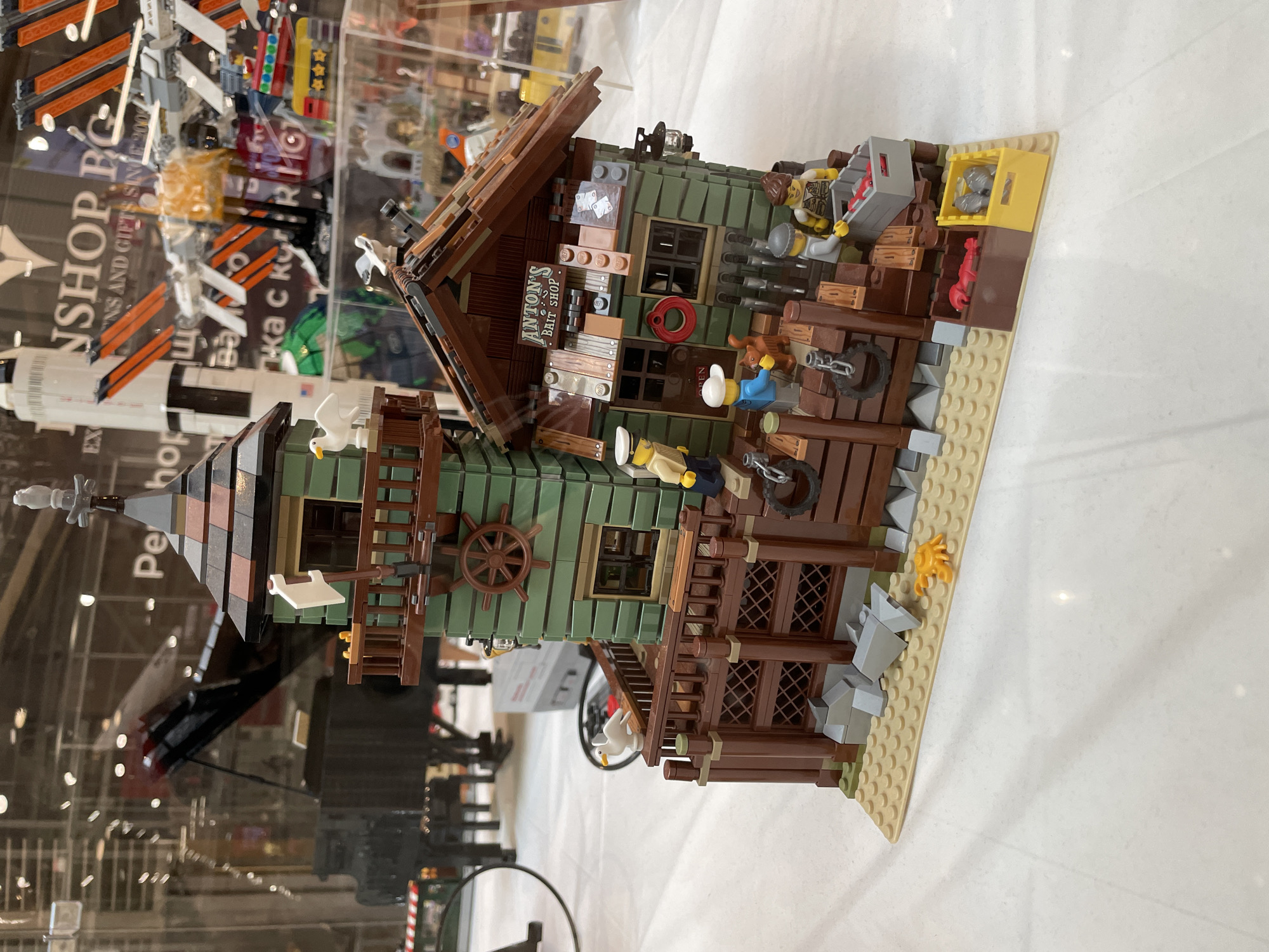 The image shows a detailed Lego model of a bait shop and lighthouse, situated on a sandy base. The bait shop is constructed with green and brown Lego bricks, featuring a sign that reads "ANTON'S BAIT SHOP." Several Lego minifigures are present, interacting with the environment. One minifigure stands on a balcony with a white bird, while another is near the shop's entrance. A minifigure in a blue shirt and white cap is interacting with a brown Lego dog. Two other minifigures are near a gray crate filled with red Lego objects, and two brown crates, one containing red Lego objects and the other gray Lego objects, are placed on the sandy base. The background, visible through a glass display, shows a grand piano, a Saturn V rocket model, and other Lego constructions. There is text in the background, including "WORKSHOP.BG" and "INS AND GIFTS SINCE 2004." The scene is clean and well-lit, suggesting it is an indoor display. The location mentioned, Sofia, Bulgaria, likely refers to the exhibition or store where this Lego model is displayed.
