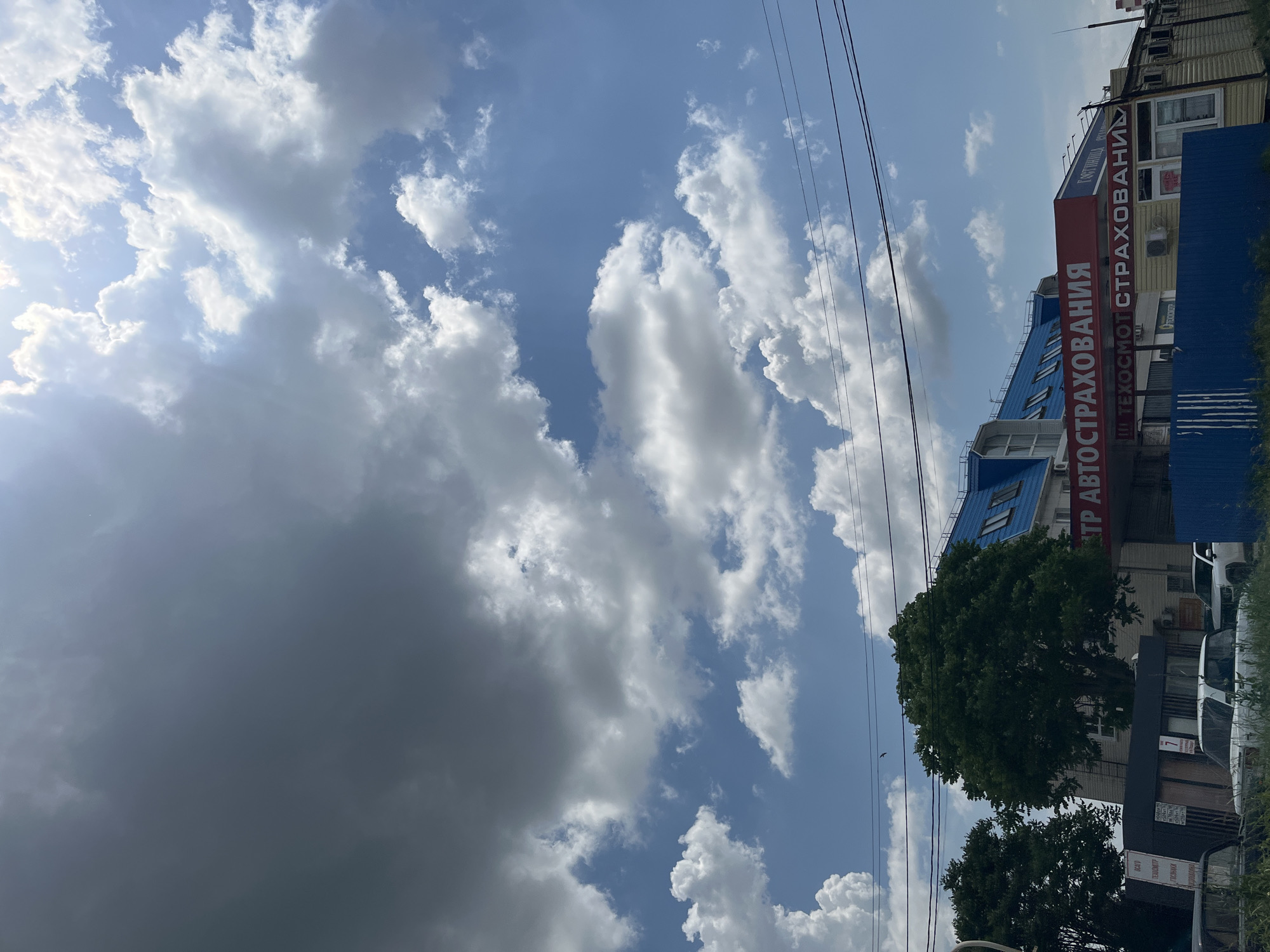 The image shows a street scene in Krasnodar, Russia. The sky is blue with scattered cumulus clouds. Overhead power lines run across the upper part of the frame. A multi-story building is visible on the right. The building is primarily blue and white with a red sign with white Cyrillic text "ЦТР АВТОСТРАХОВАНИЯ Ш ТЕХОСМОТ СТРАХОВАНИЯ". Trees partially obscure the lower floors of the building. Cars are parked in front of the building. The image is rotated 90 degrees clockwise.
