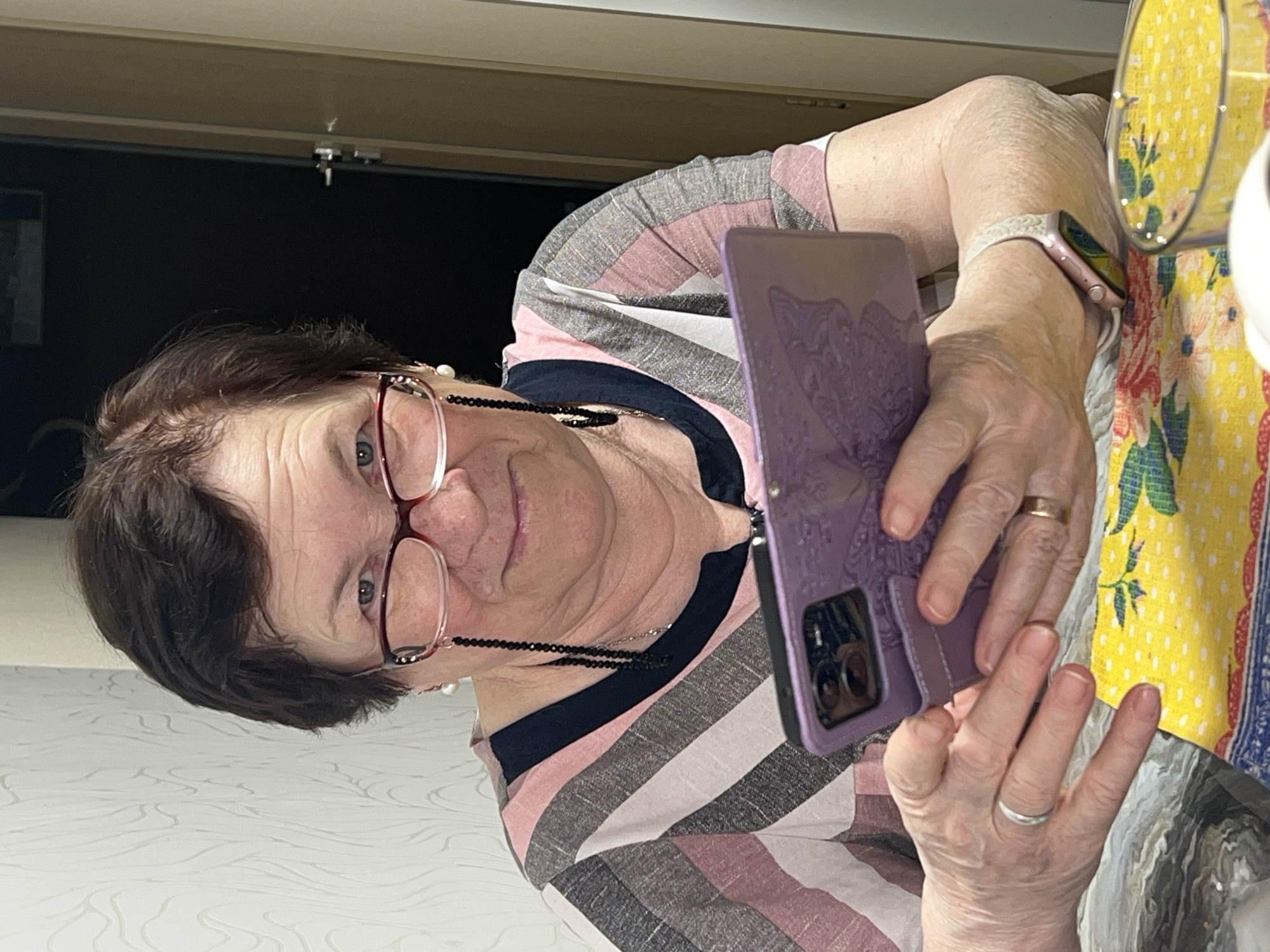 An older woman is depicted in a close-up, indoor shot, likely taken as a selfie or during a video call in Perm, Russia. She has short, dark brown hair and wears reddish-brown framed glasses, which are connected by a black beaded and silver chain around her neck. Her expression is neutral, with her eyes looking towards the camera lens.
She is wearing a horizontally striped shirt with alternating bands of pink, grey, white, and black. On her left wrist, she sports a light pink smart watch with a matching band. She holds a smartphone encased in a purple, textured wallet-style case, which appears to have an embossed design. A gold-colored ring is visible on her right ring finger, and a silver-colored ring is on her left pinky finger.
The background behind her is dark, suggesting an interior space like a room or doorway. To her right, part of a yellow textile with a white dot pattern and a floral design is visible, possibly a tablecloth or placemat. The image itself is significantly tilted, reflecting the angle at which it was captured. No discernible text is visible within the frame.