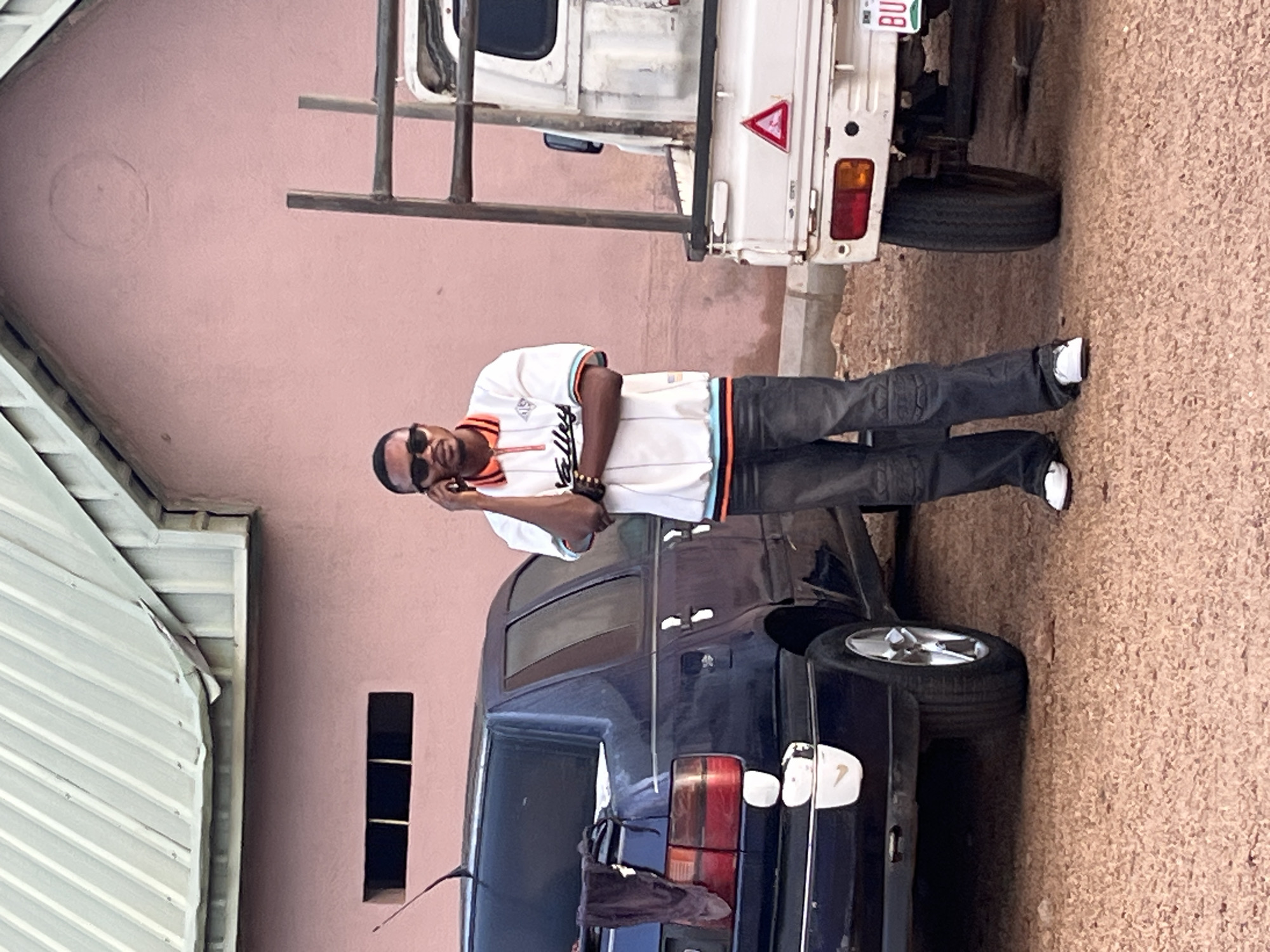 A man stands between a dark blue car and a white mini truck in an outdoor setting. The man is wearing sunglasses, a white jersey with orange and black trim, dark jeans, and white sneakers. He is looking at the camera while holding a mobile phone to his ear. The dark blue car has its trunk partially open. The white mini truck has a ladder rack attached to the back. The background includes a pale pink wall with small, rectangular cutouts and a corrugated metal structure. The ground is covered with fine gravel. The location is presumed to be in Jos, Nigeria.
