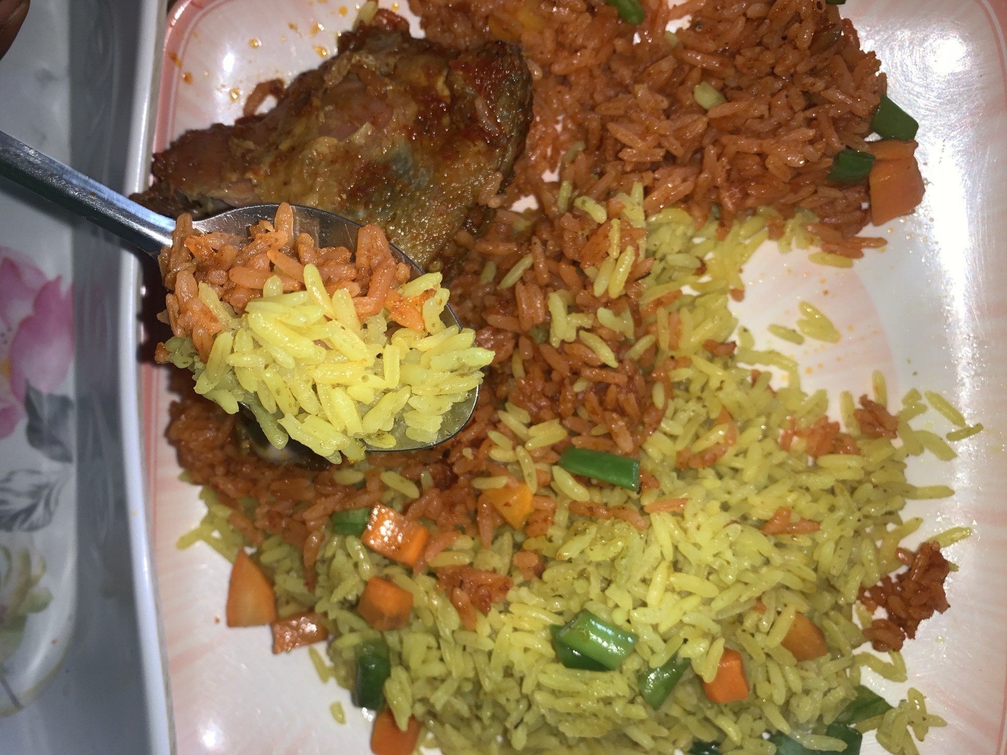 A top-down, close-up shot shows a plate of food, possibly in Kabong, Nigeria. The plate holds two kinds of rice: yellow rice with diced carrots and green beans, and red rice. A piece of fried fish is next to the red rice. A spoonful of mixed yellow and red rice is raised above the plate. The plate is white with a pink floral border. The surface beneath the plate is light colored.