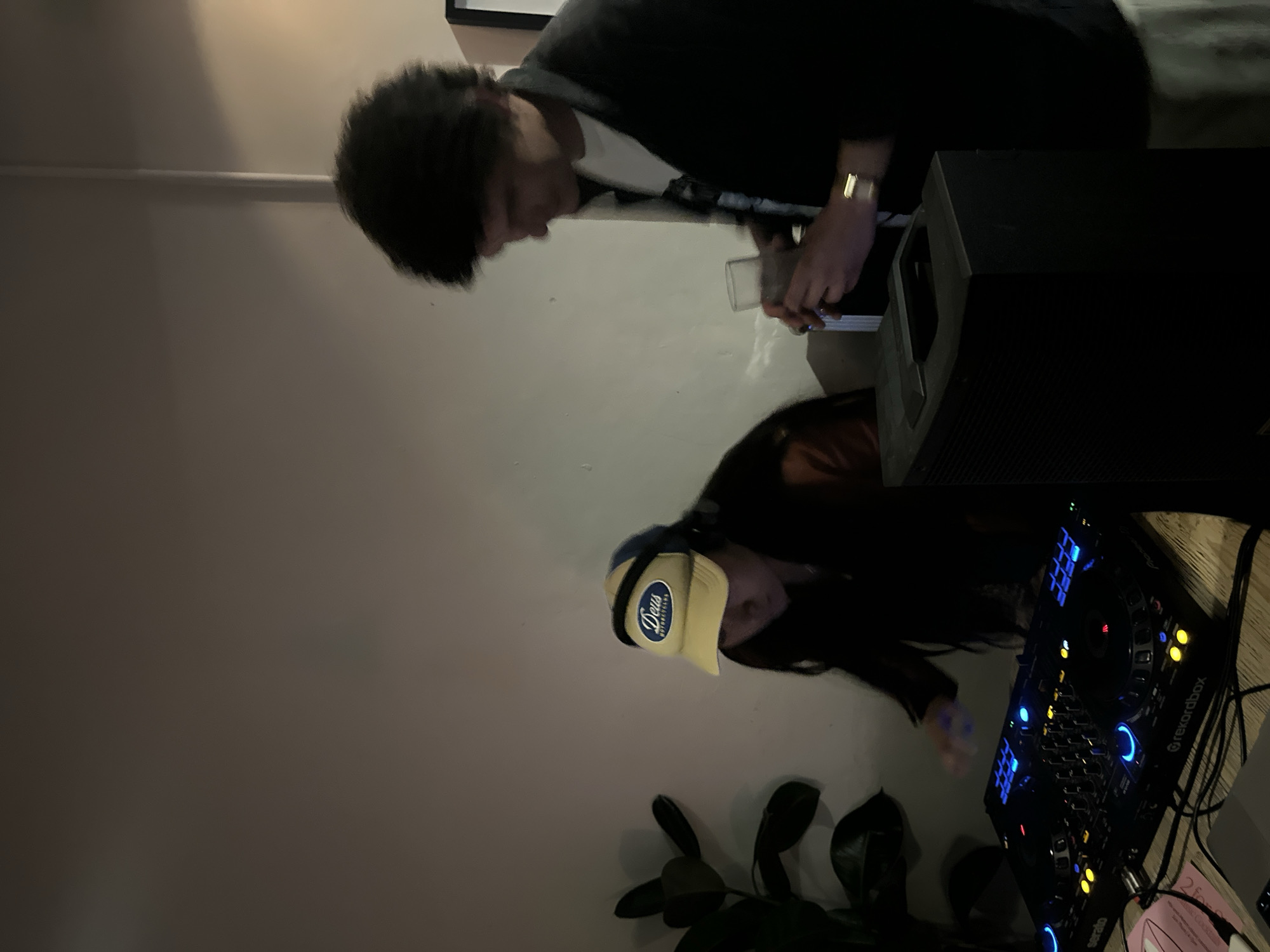 Two people are near a DJ setup. One person, wearing a black jacket and a gold watch, is holding a small electronic device and looking at it. The second person is wearing a hat with "Deus Ex Machina" written on it and is operating a DJ mixer. A large black speaker is to the right of the first person. A potted plant is near the DJ setup. The background is a light-colored wall. It is not possible to confirm the location is London, United Kingdom.