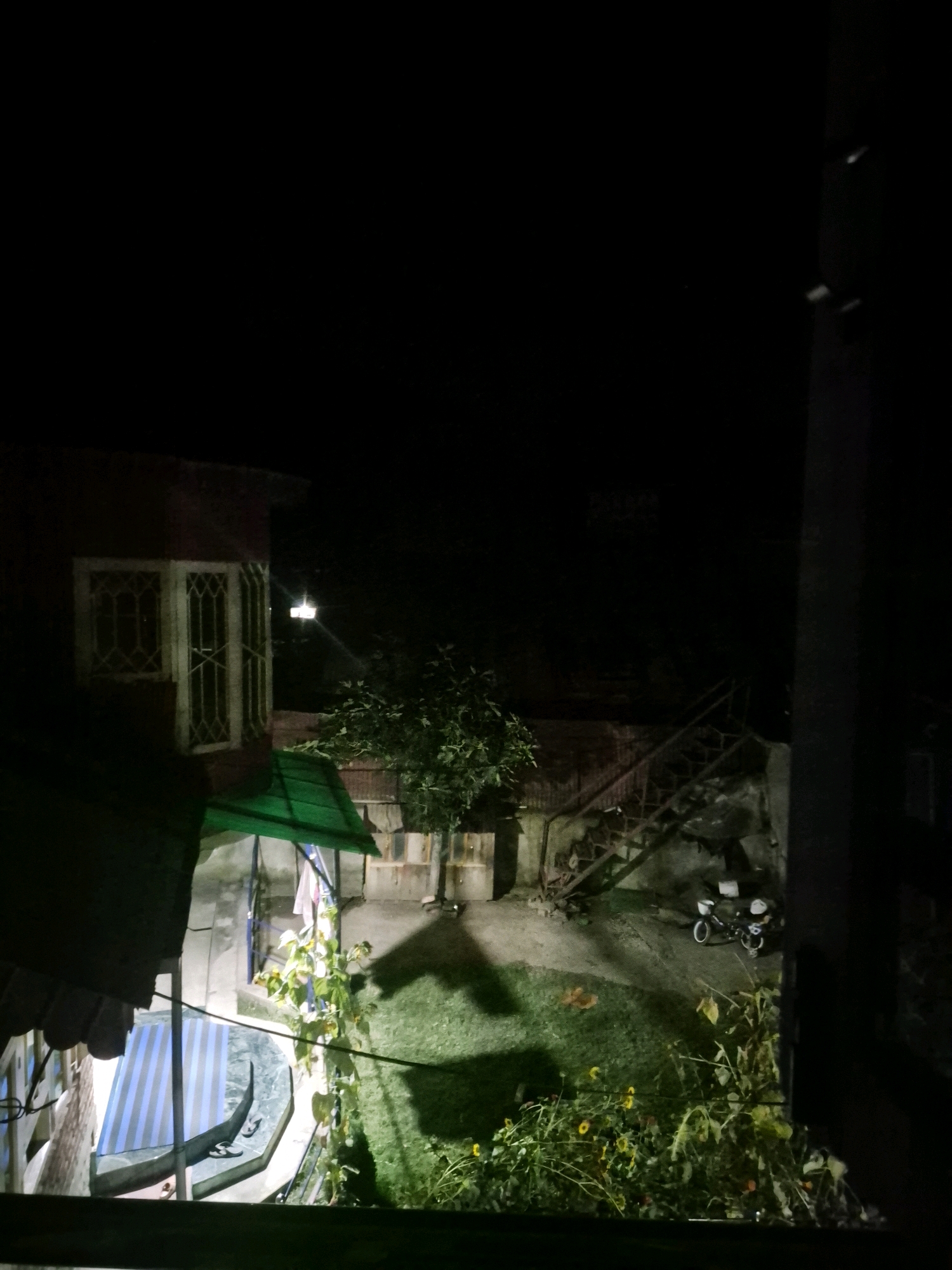 The image depicts an outdoor courtyard at night, illuminated by artificial lights. On the left, a portion of a building is visible with a red brick facade and a window featuring a grid pattern. A green awning extends from the building. In the center of the courtyard, a tree casts a shadow on the grass. To the right, a set of outdoor stairs leads upwards, and a parked motorcycle is visible at the base of the stairs. The ground is covered in grass and some plants. The overall scene suggests a residential or semi-residential area in Bandipora, India, likely a backyard or patio.