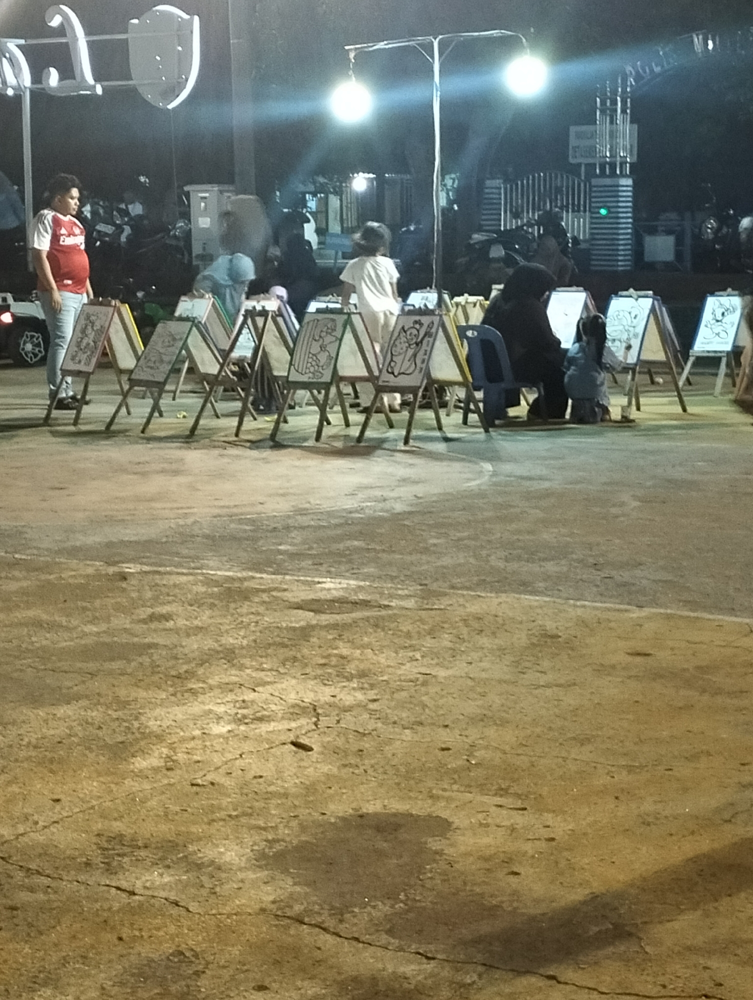 The image depicts an outdoor event at night in Lhokseumawe, Indonesia. A series of small easels with coloring pages face away from the camera, forming a semi-circle. Some children and adults are sitting at the easels, appearing to be engaged in coloring. A man wearing a red shirt and jeans stands to the side, observing the scene. In the background, there are parked motorbikes, a light-colored structure, and a metal gate with the words "Rock's Music" visible. Two bright lights are elevated above the scene, illuminating the area. The ground is a light-colored pavement with visible cracks.