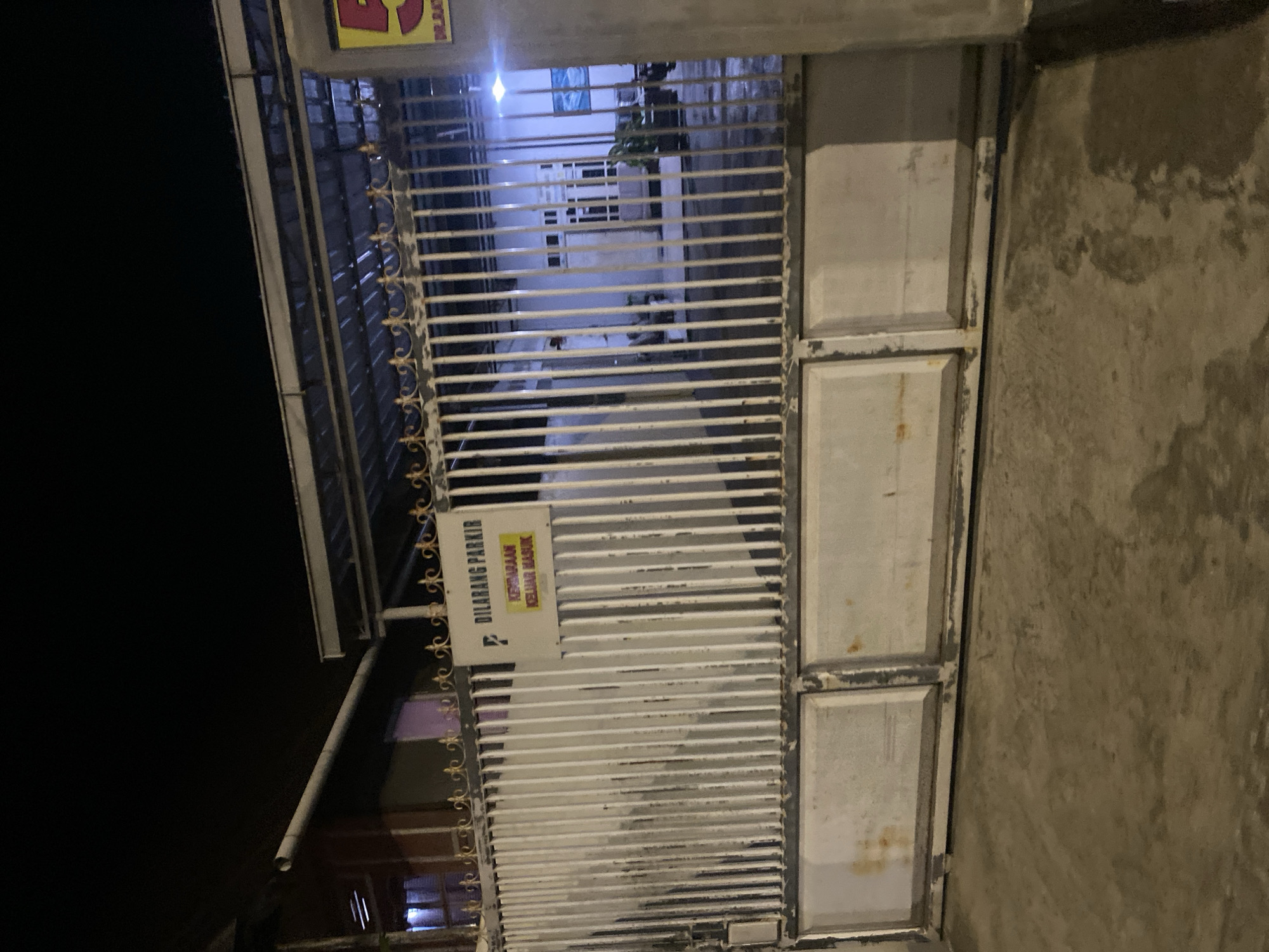 The image shows a metal gate in front of a building at night. There is a sign on the gate that says "DILARANG PARKIR KENDARAAN KELUAR MASUK" which translates to "NO PARKING VEHICLE IN AND OUT." The building appears to be a home or a small business, with windows and lights visible from the outside. The area is paved and appears to be a residential or commercial street. The location is in Bantaeng, Indonesia.