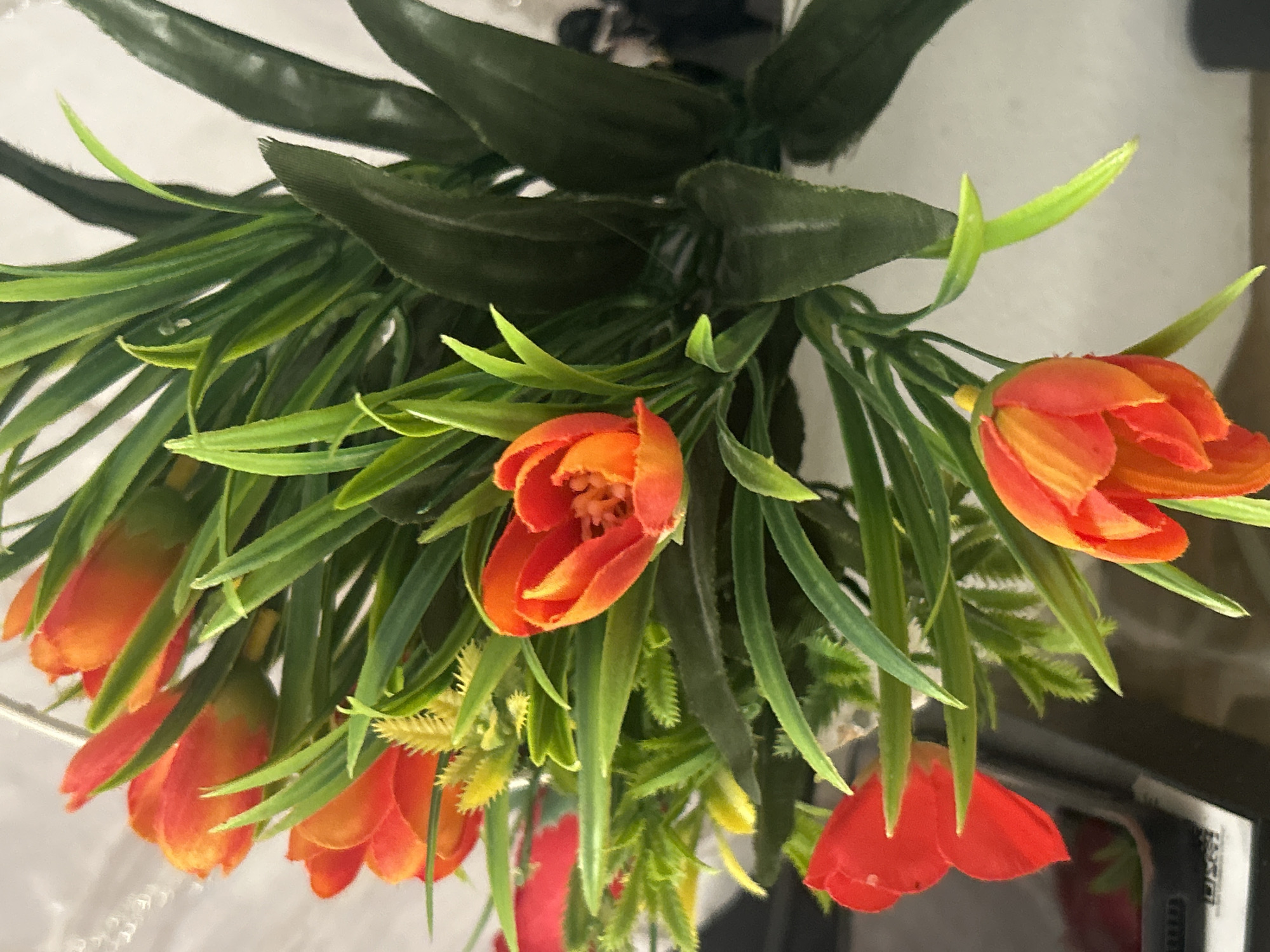 The image shows a close-up of a bouquet of artificial tulips with vibrant orange and red petals and green leaves. The flowers are arranged in a way that suggests they are part of a larger display, possibly on a table or in a vase. The background is blurred and light-colored, with some hints of darker shapes that could be furniture or other objects. There is no indication of the city of Georgiyevskaya, Russia in the image, nor any people or actions.