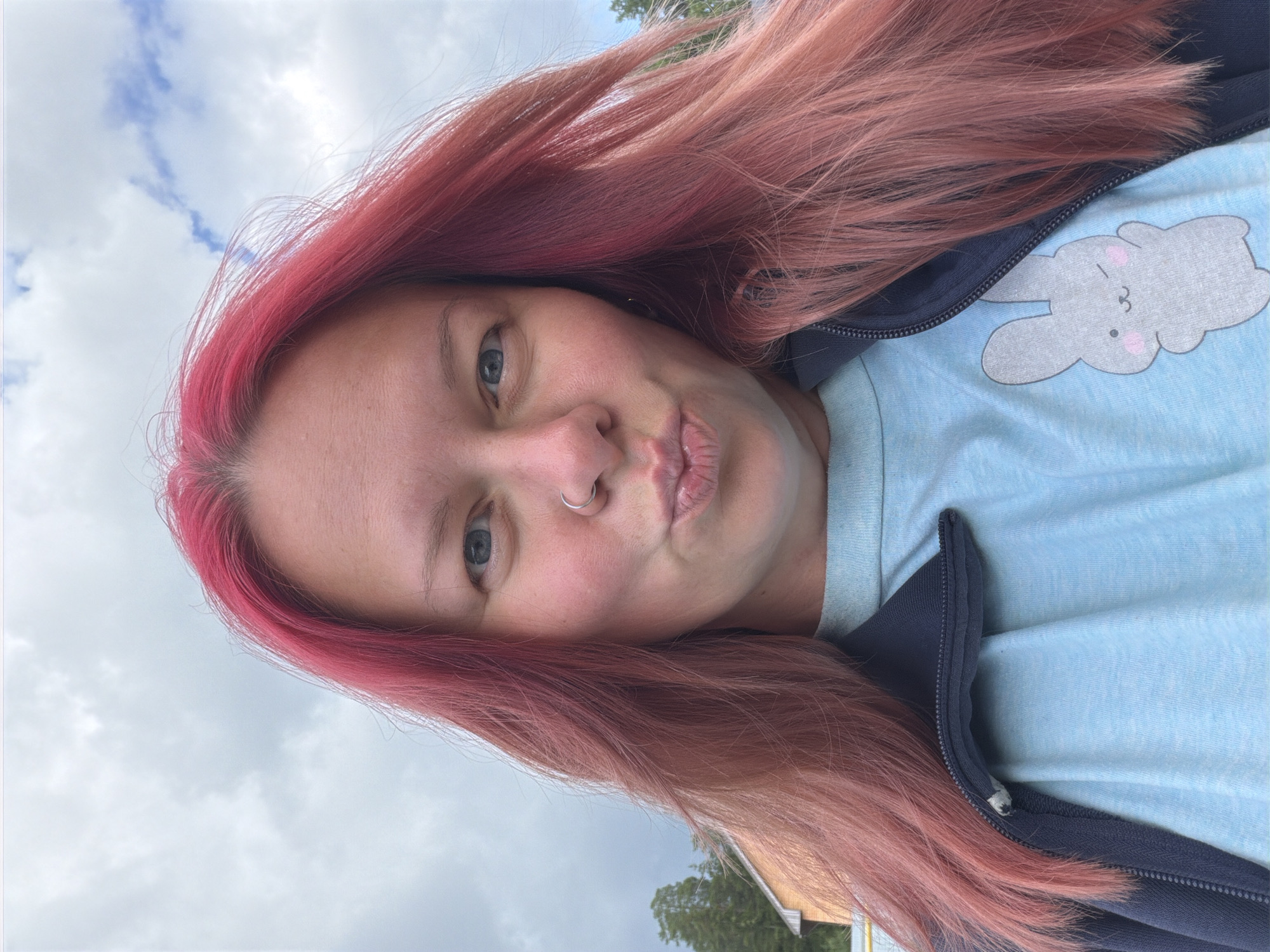 A woman with pink hair is taking a selfie outdoors. She is wearing a light blue t-shirt with a bunny graphic and a dark blue zip-up jacket. Her lips are pursed, and she has a nose ring. The background shows a cloudy sky and some trees and a building, possibly in Chaikovsky, Russia.