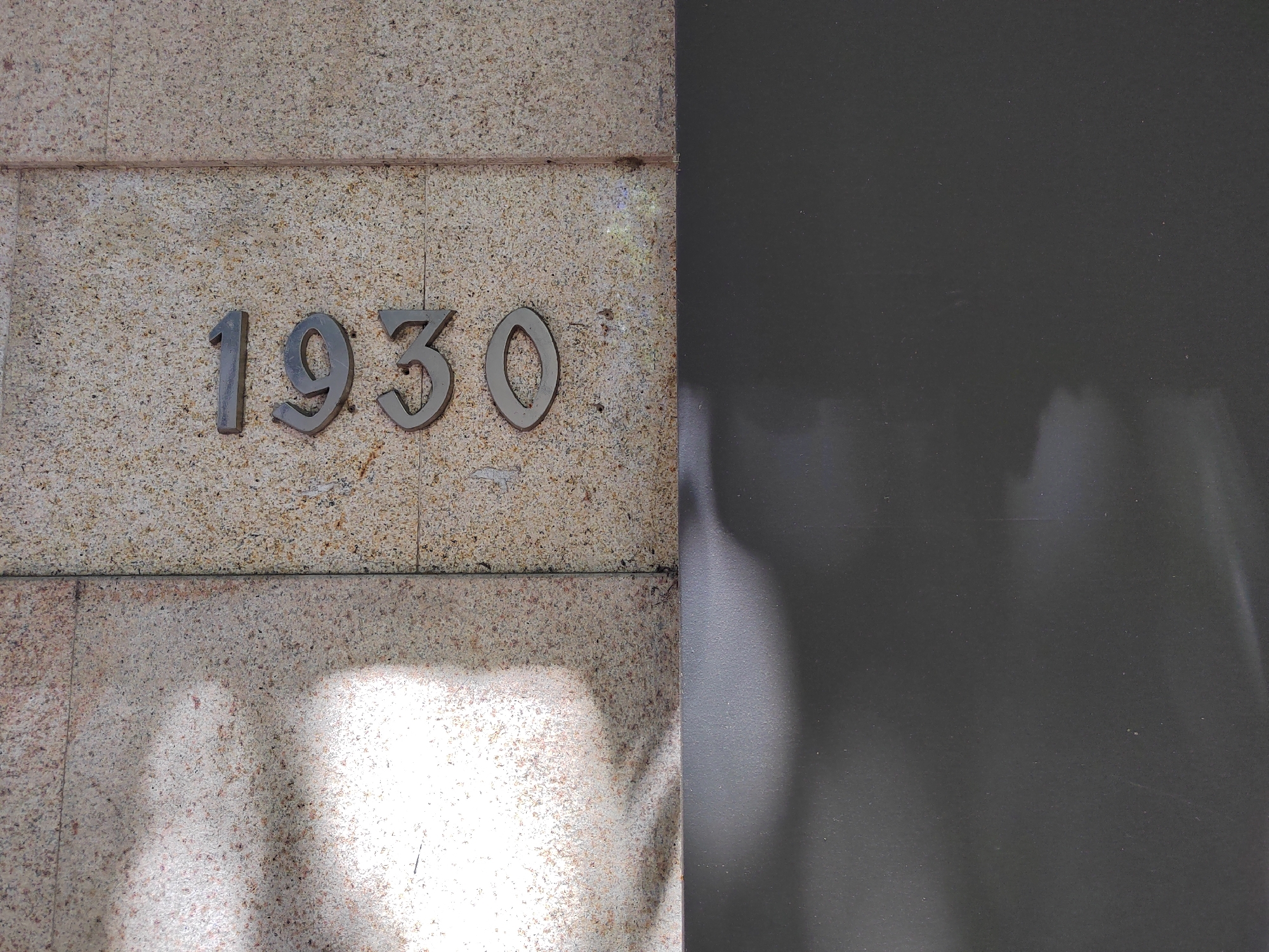 A building in Podgorica, Montenegro, displays the number 1930 on a textured stone façade. The numbers are made of a metallic material, possibly bronze or a similar alloy. The stone has a mottled appearance with variations in color from light to dark. To the right of the stone façade, there is a plain, dark-colored surface. The lower part of the stone façade is illuminated by sunlight.