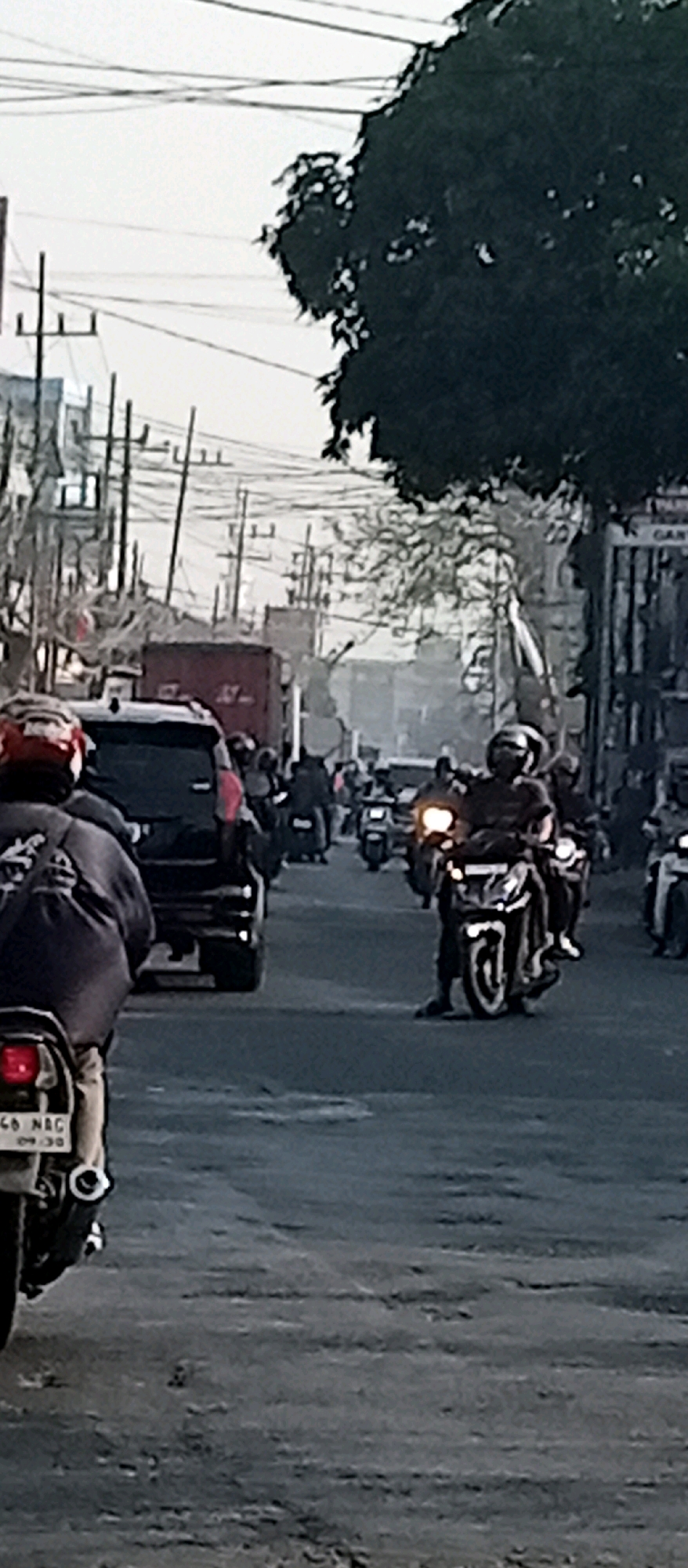 The image depicts a street scene in Wadungasri, Indonesia, characterized by heavy traffic dominated by motorcycles. In the foreground, a person wearing a red helmet and a dark jacket is riding a motorcycle. Behind them, a black SUV is visible. The middle ground features a convoy of motorcycles, with riders wearing helmets and casual clothing. One motorcycle's headlight is illuminated, indicating movement. In the background, utility poles with numerous wires stretch across the sky, a common sight in urban Indonesian settings. Buildings line the street, and a large, dark tree with dense foliage partially obscures the upper right portion of the image. The road surface appears to be asphalt, with some texture suggesting wear. The overall atmosphere suggests a busy street during daylight hours.